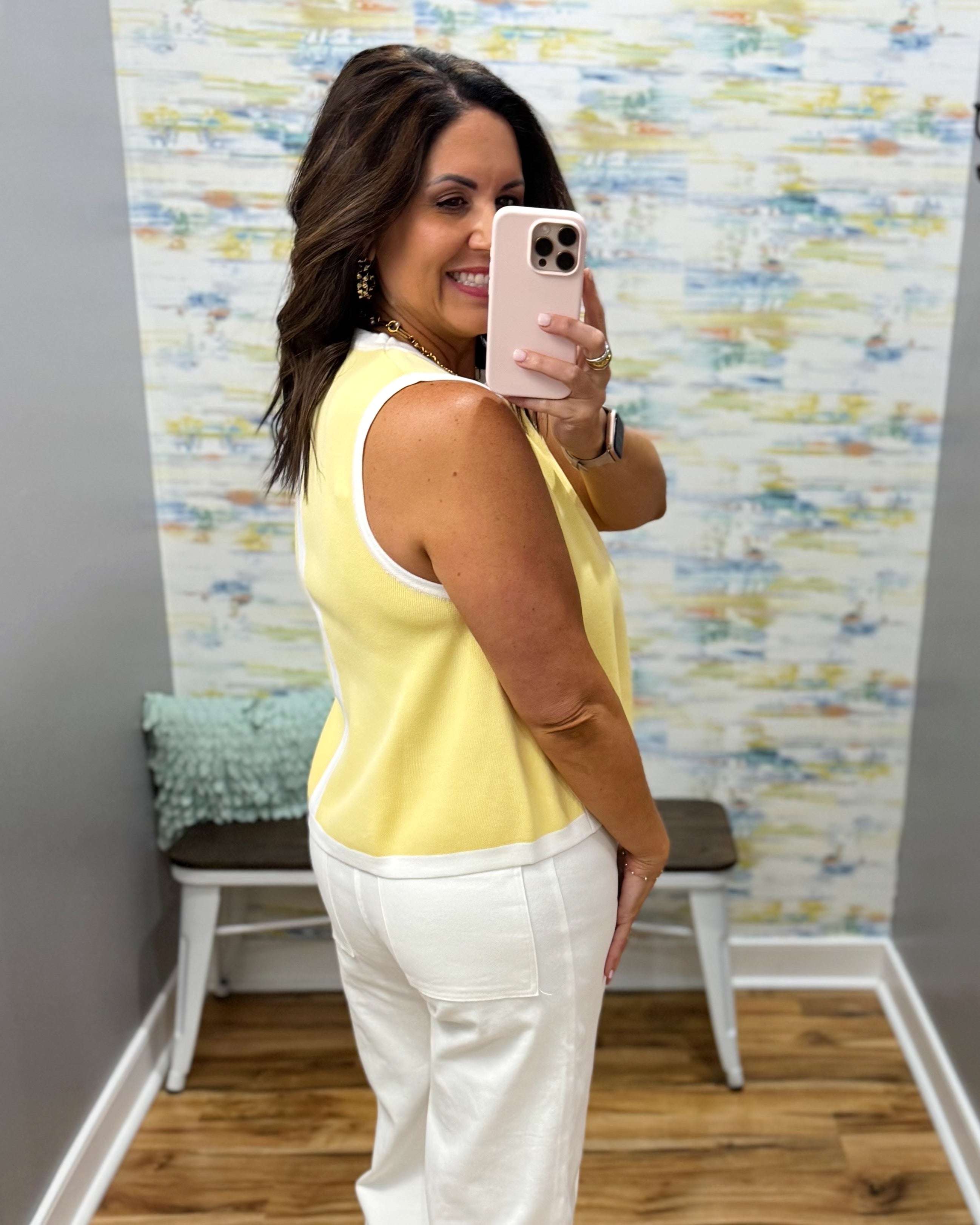Woman taking a mirror selfie wearing a yellow top and white pants in a room with colorful wall art.