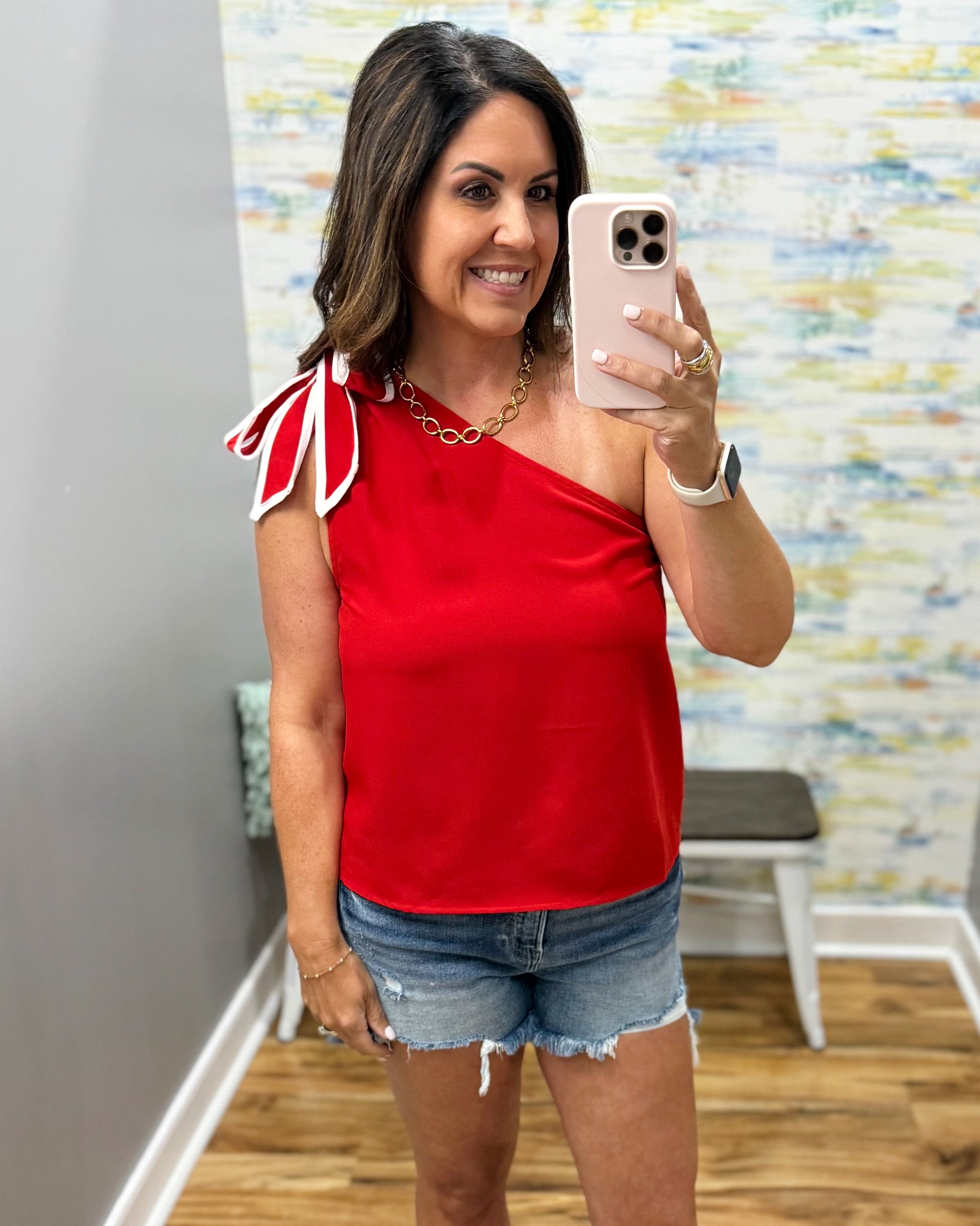 Woman taking a mirror selfie wearing a red one-shoulder top and denim shorts.