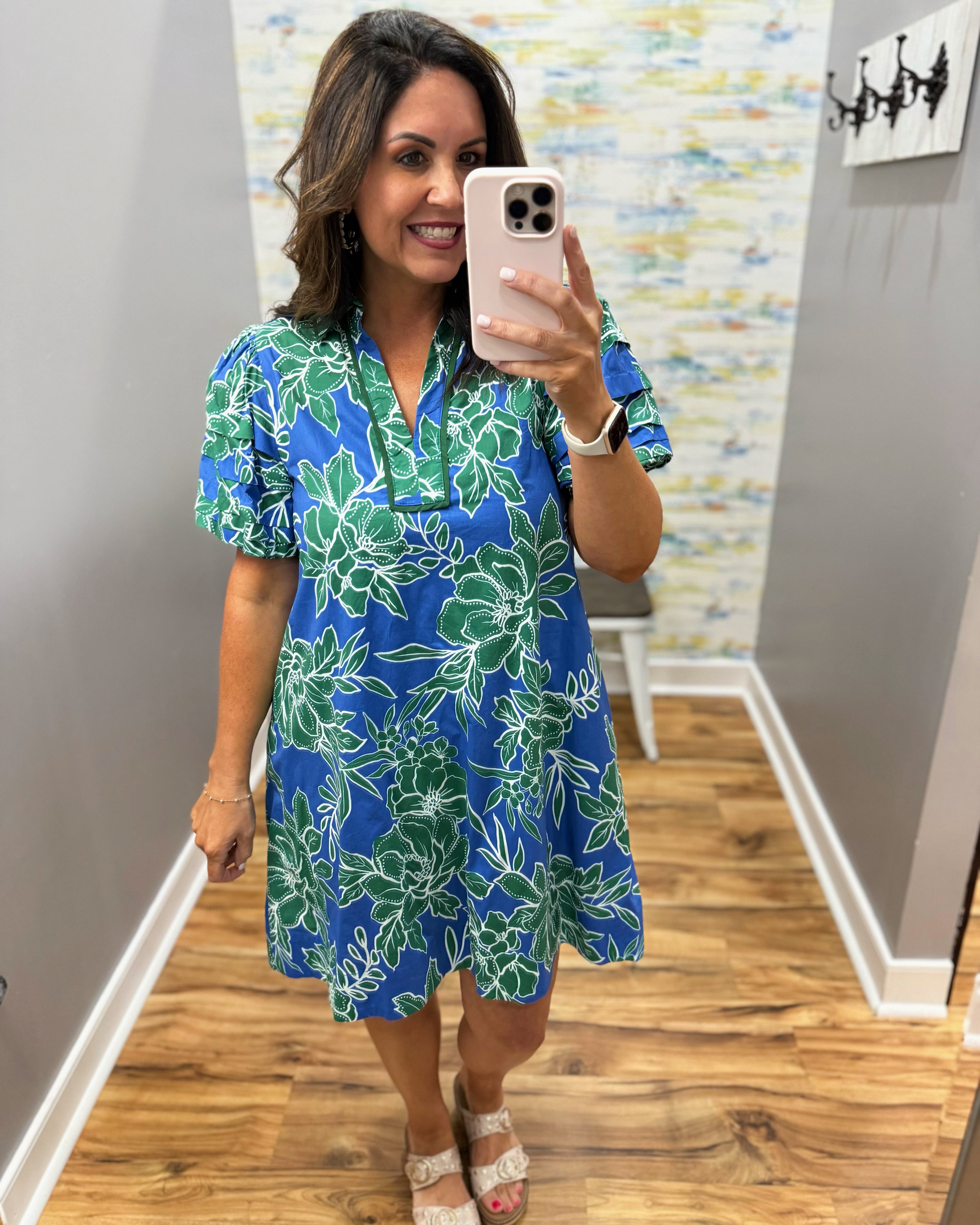 Woman taking a mirror selfie wearing a blue floral dress in a room with wooden flooring and a colorful wall.