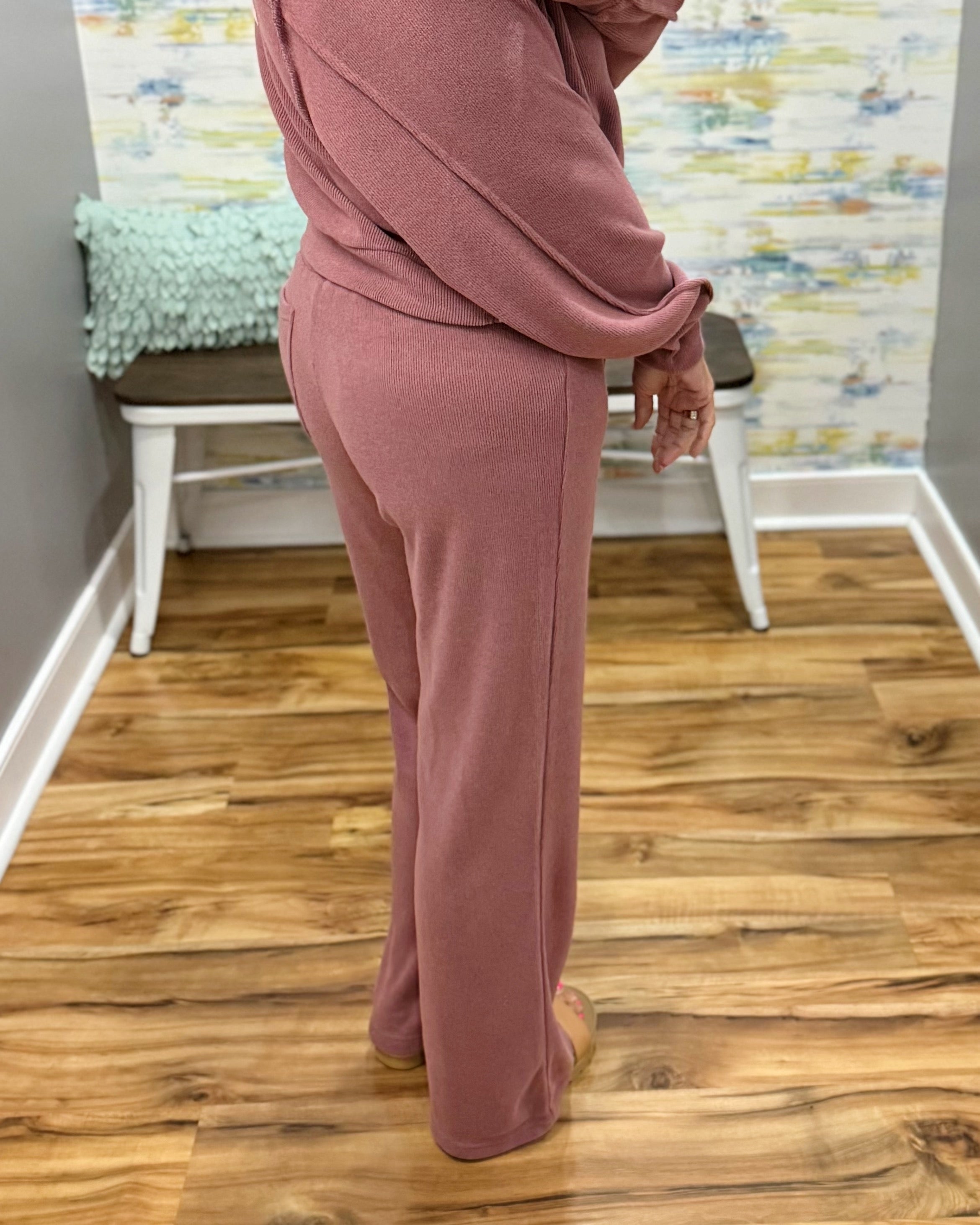 A person wearing a dusty rose pink two-piece lounge set consisting of a long-sleeve top and matching wide-leg pants, standing on a wood floor in front of a white bench.