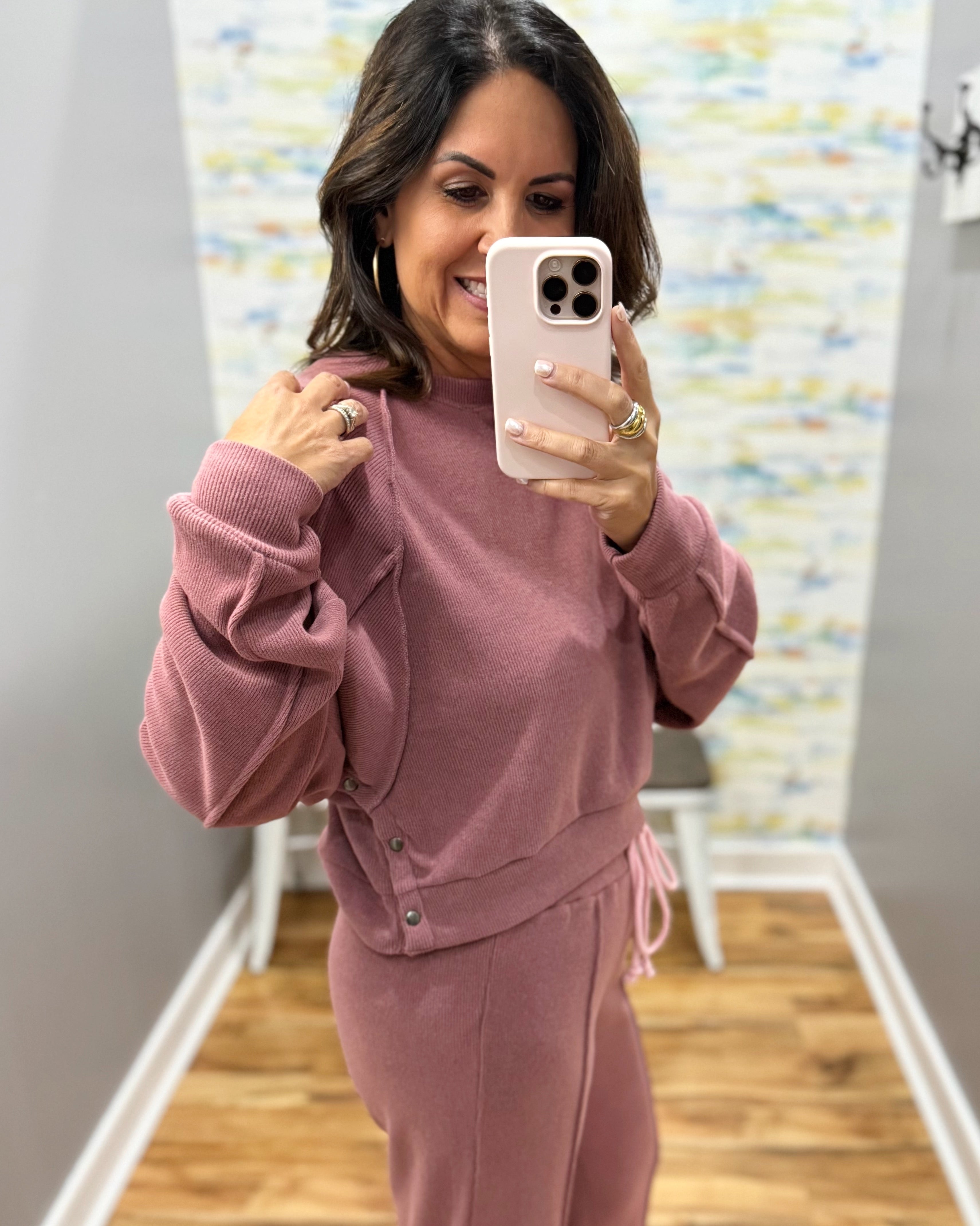 A woman takes a mirror selfie wearing a dusty rose pink two-piece lounge set consisting of a cropped sweatshirt with gathered sleeves and matching jogger pants. She is holding a light pink phone in a home setting with a wood floor and patterned wallpaper.