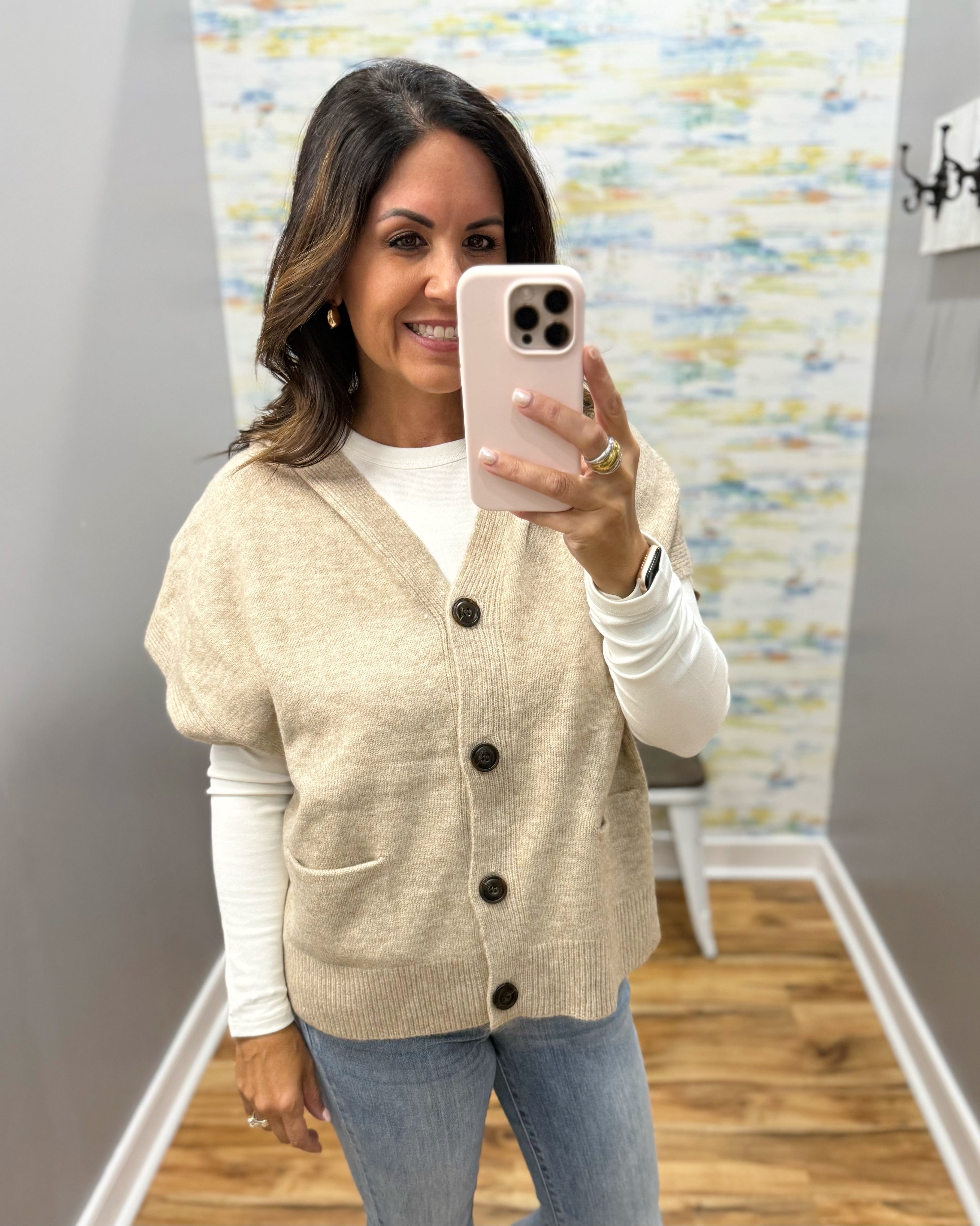 A woman takes a mirror selfie wearing a beige knit cardigan with black buttons over a white long-sleeve shirt and blue jeans.