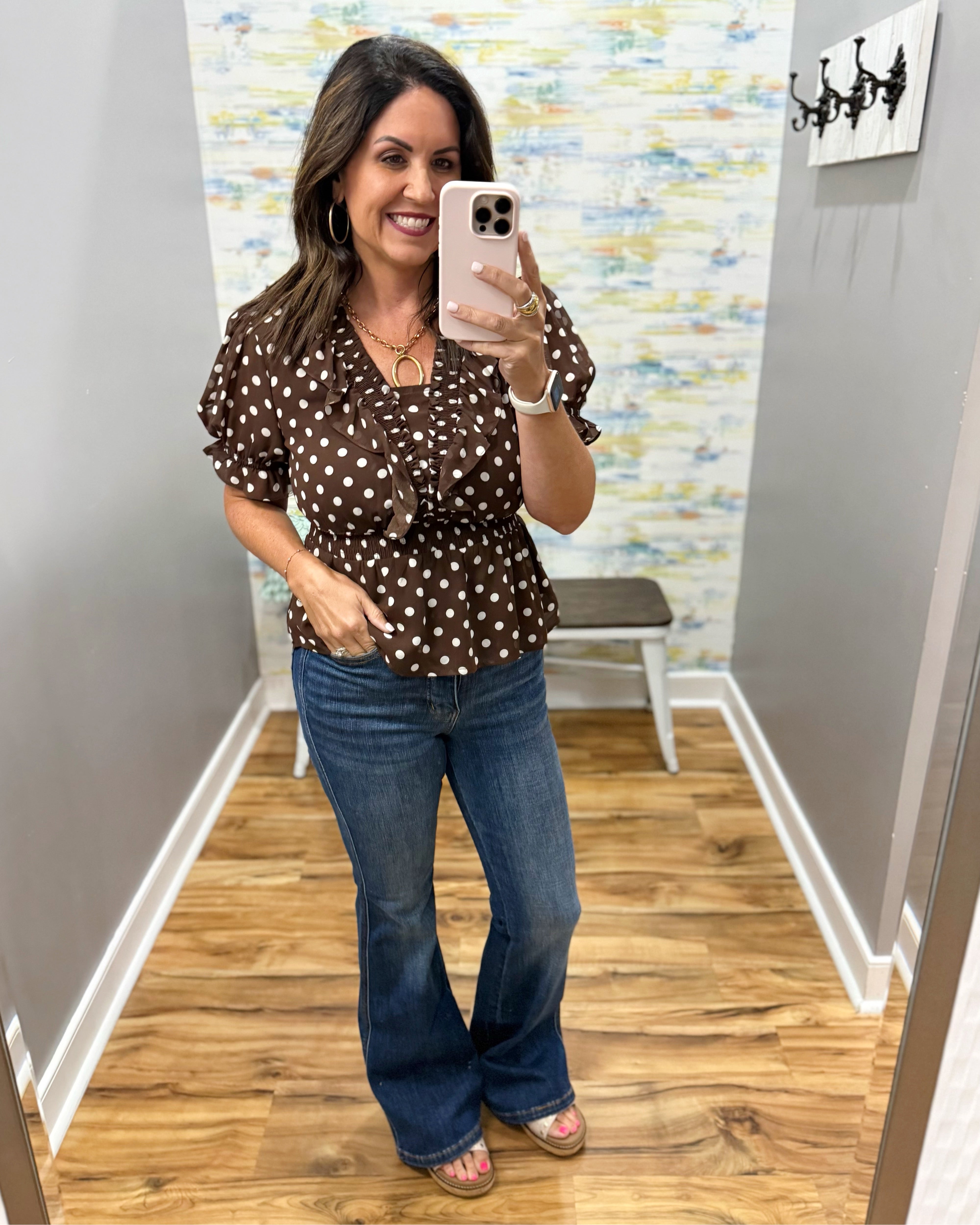 A woman takes a mirror selfie in a fitting room wearing a brown short-sleeve blouse with white polka dots and blue flare jeans.
