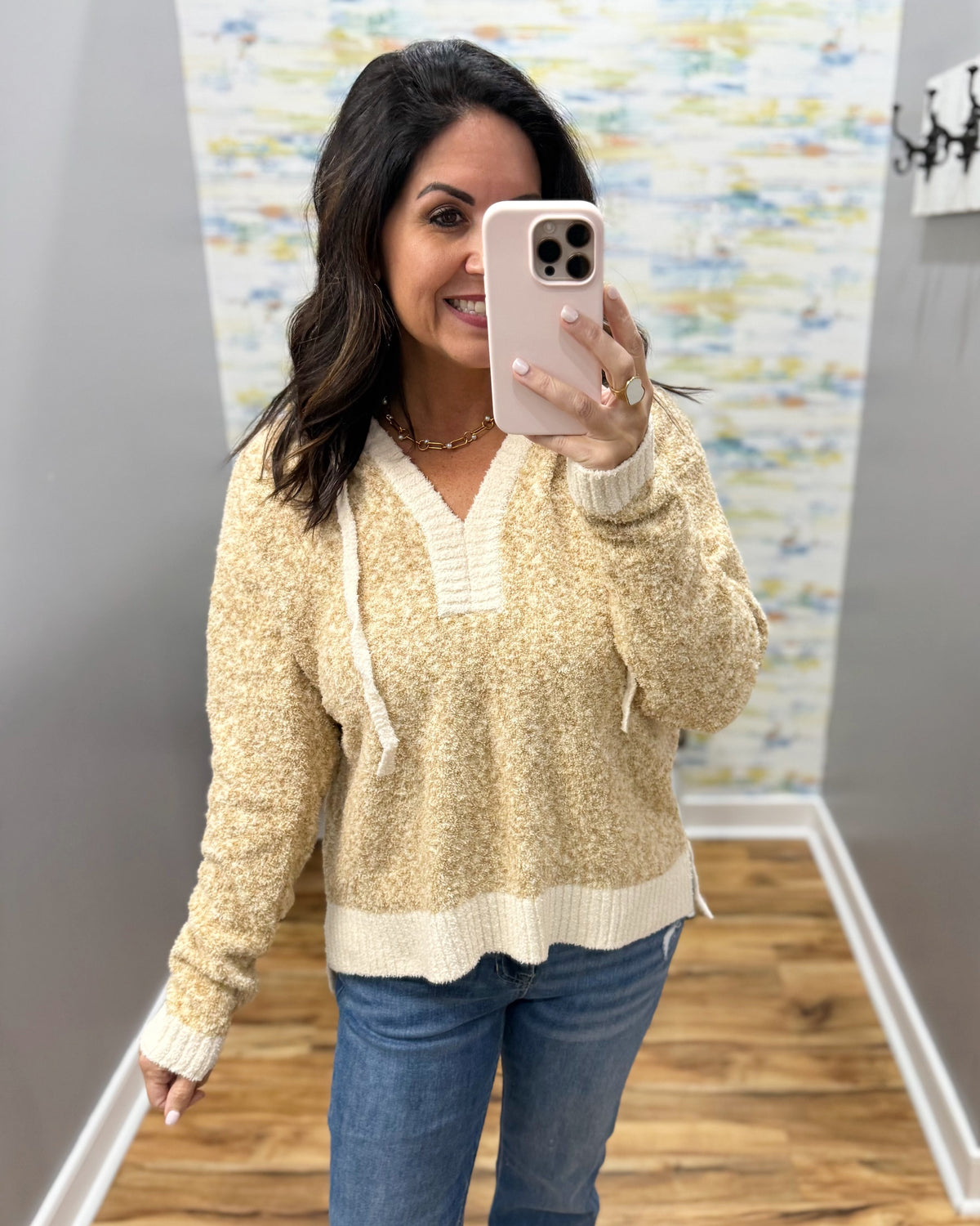 A woman takes a mirror selfie wearing a cream and beige marled knit V-neck sweater with white drawstrings, paired with blue jeans, in an indoor hallway.