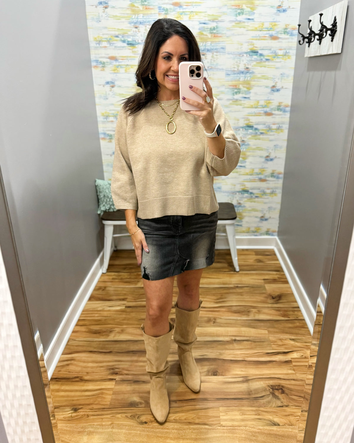 A woman takes a mirror selfie in a fitting room wearing a beige knit crewneck sweater, a black denim mini skirt, and beige knee-high boots.