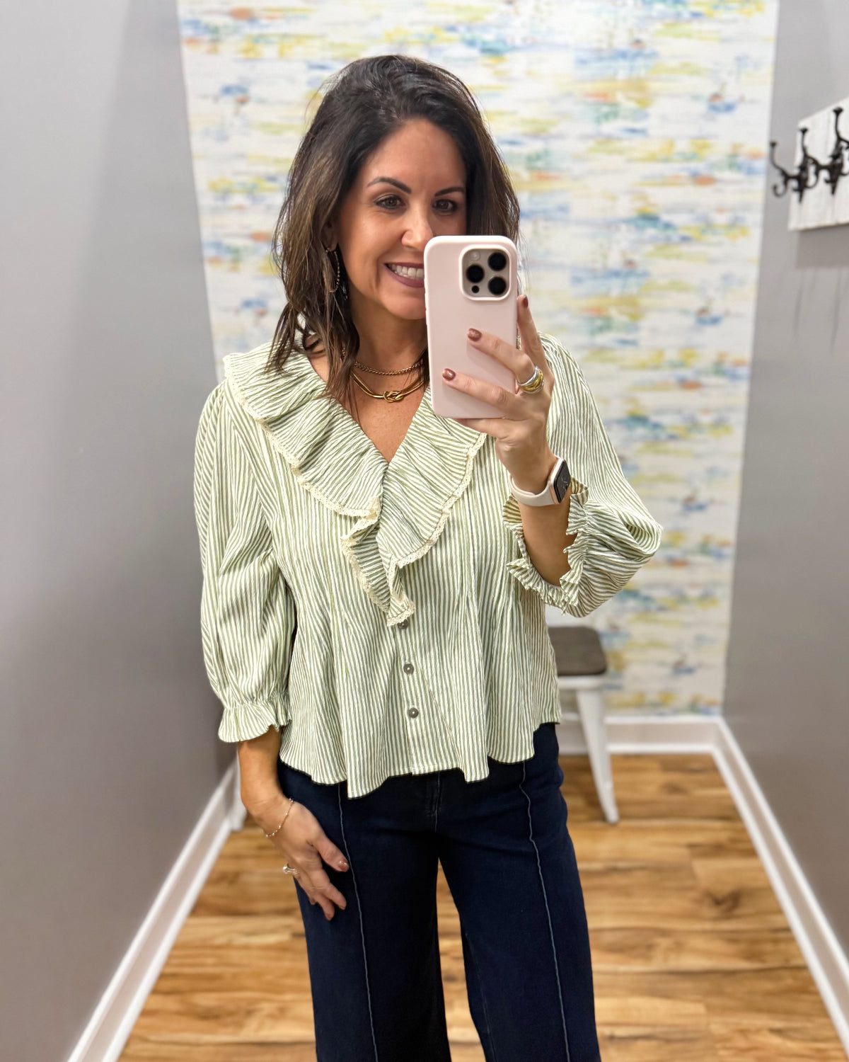 A woman takes a mirror selfie wearing a light green and white vertically striped blouse with ruffle details and dark blue jeans in an indoor boutique setting.