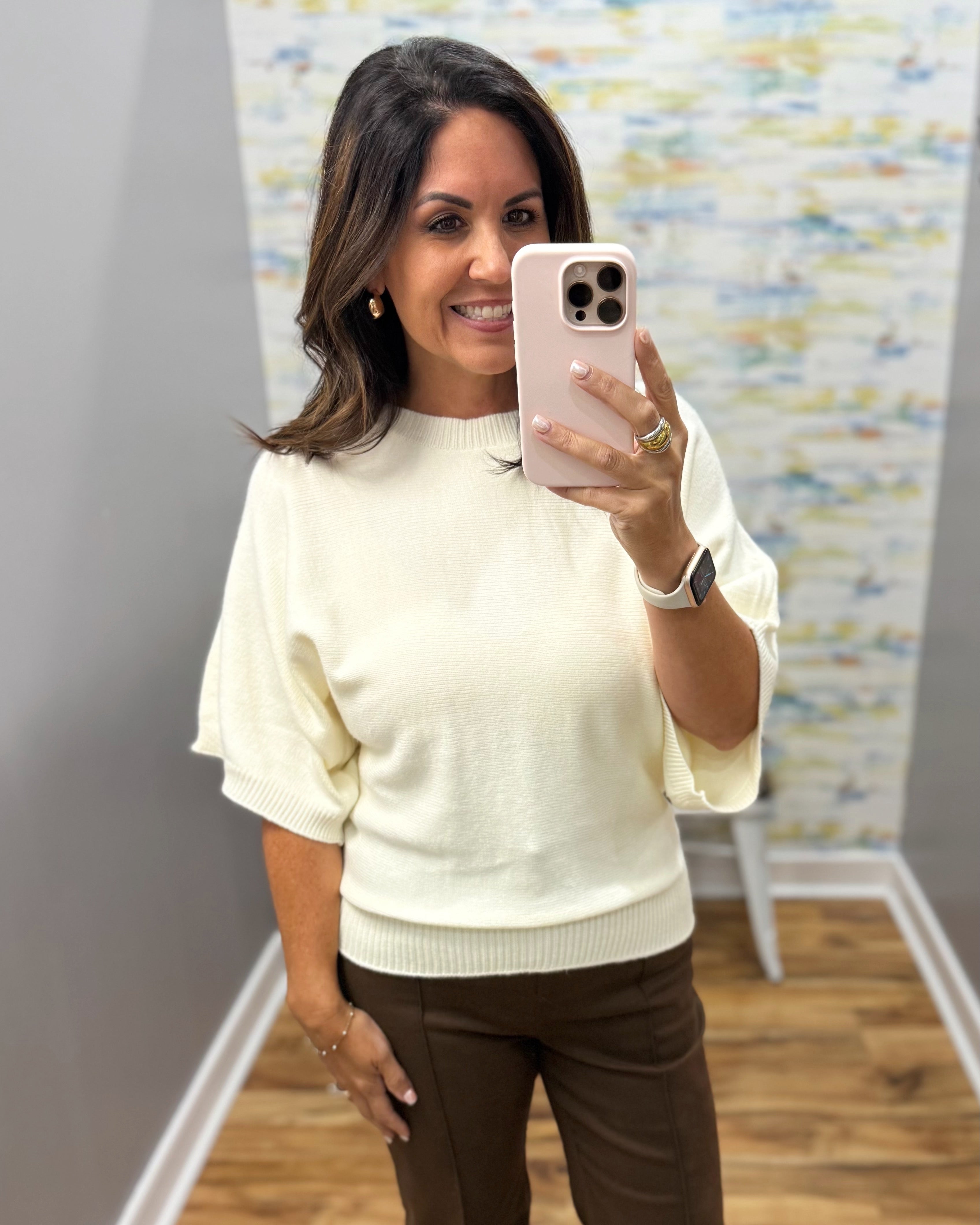 A woman taking a mirror selfie in a boutique, wearing a cream-colored short-sleeve knit sweater and brown pants.