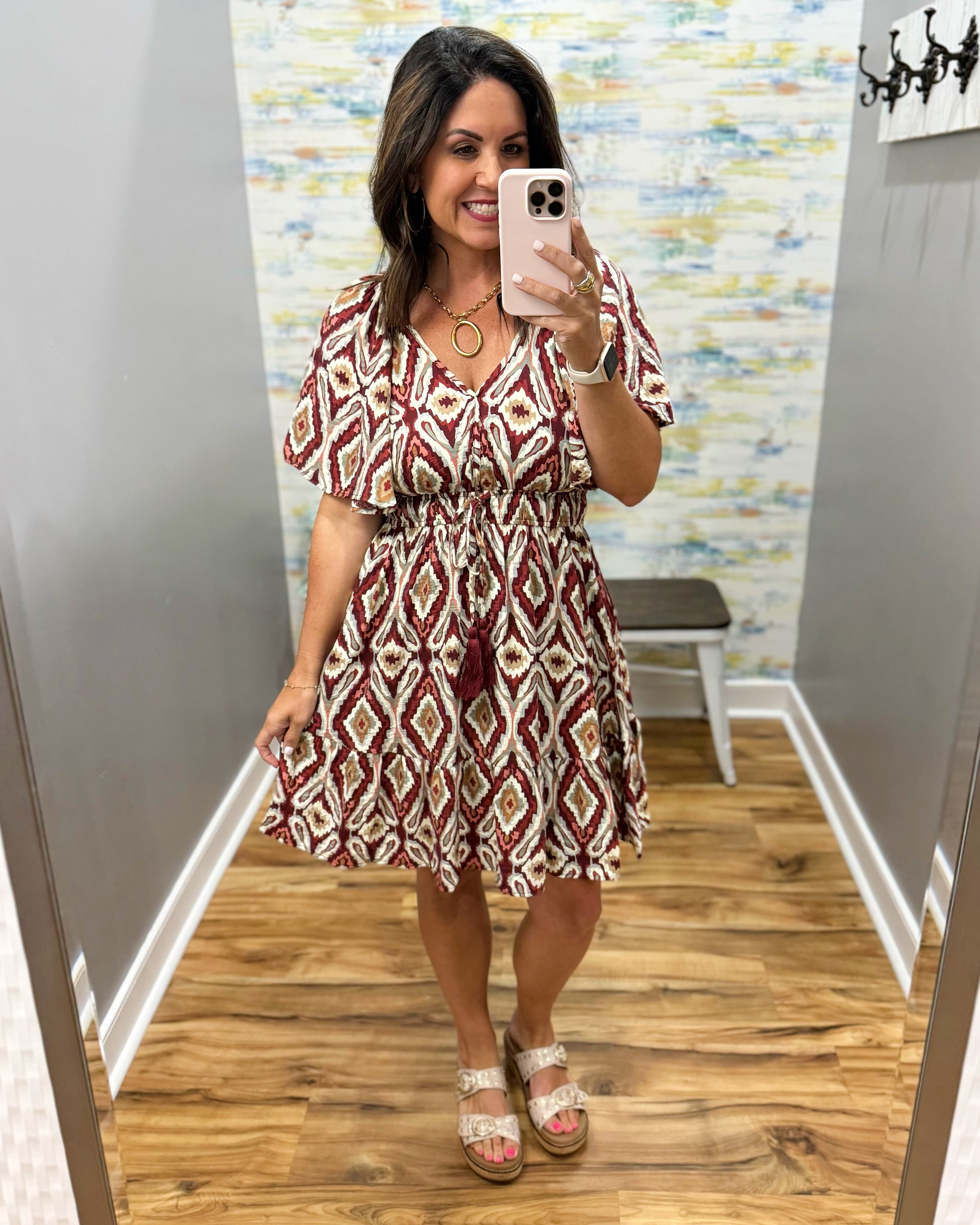 A woman takes a mirror selfie in a fitting room wearing a short-sleeve V-neck dress with a red, beige, and white geometric diamond pattern. She is wearing beige platform sandals and gold jewelry.