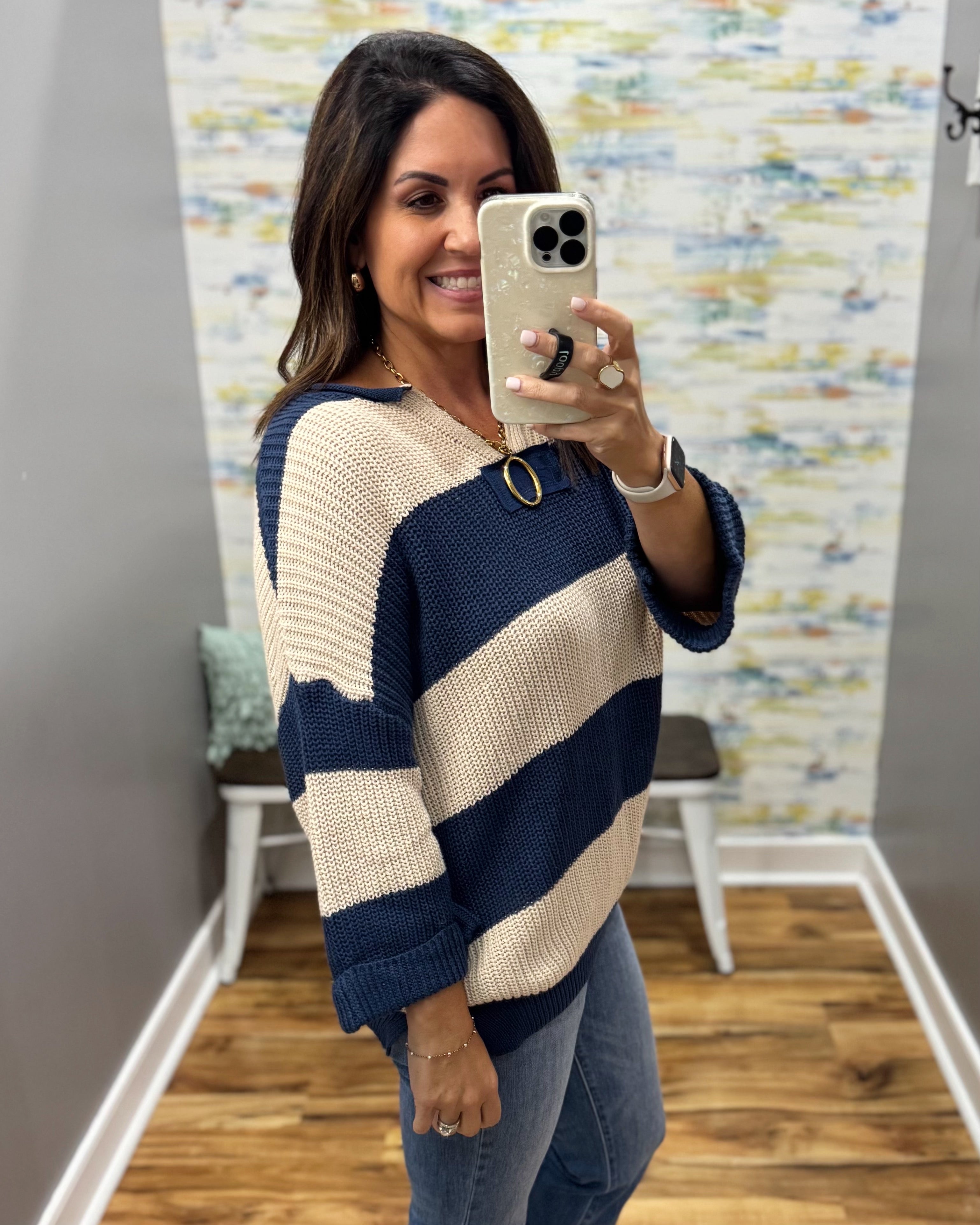 A woman takes a mirror selfie wearing a knit sweater with bold navy blue and cream horizontal stripes, paired with blue jeans.