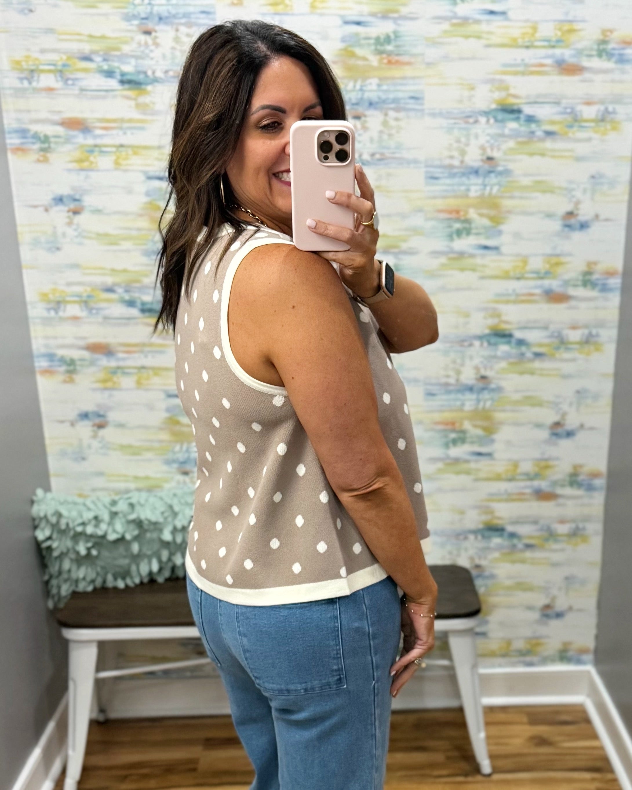 Woman taking a mirror selfie wearing a polka dot top and jeans in a room with colorful wall art.