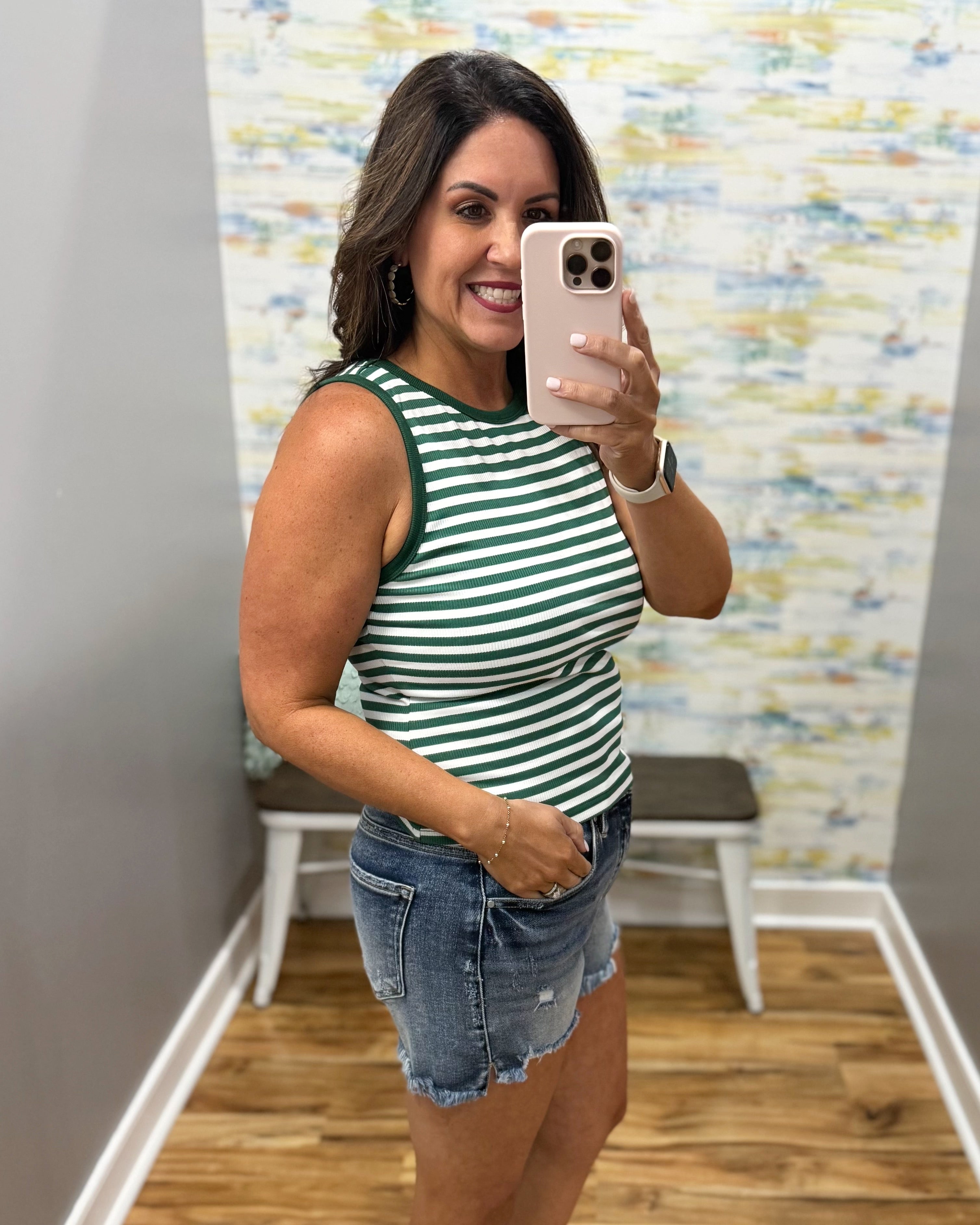Woman taking a mirror selfie wearing a green and white striped tank top and denim shorts.