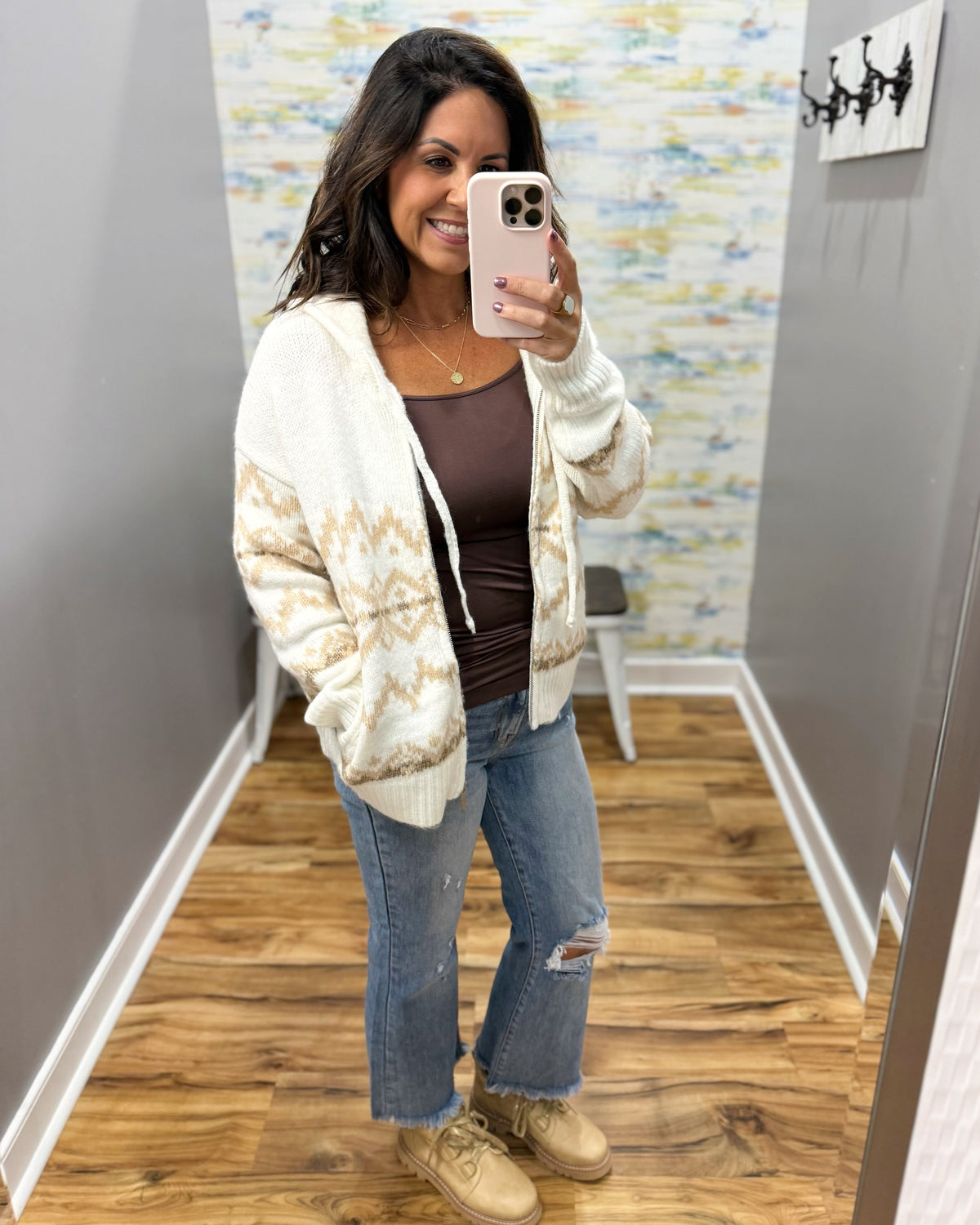 A woman takes a mirror selfie in a fitting room wearing a cream and tan patterned knit zip-up hoodie over a brown top, blue jeans, and beige sneakers.