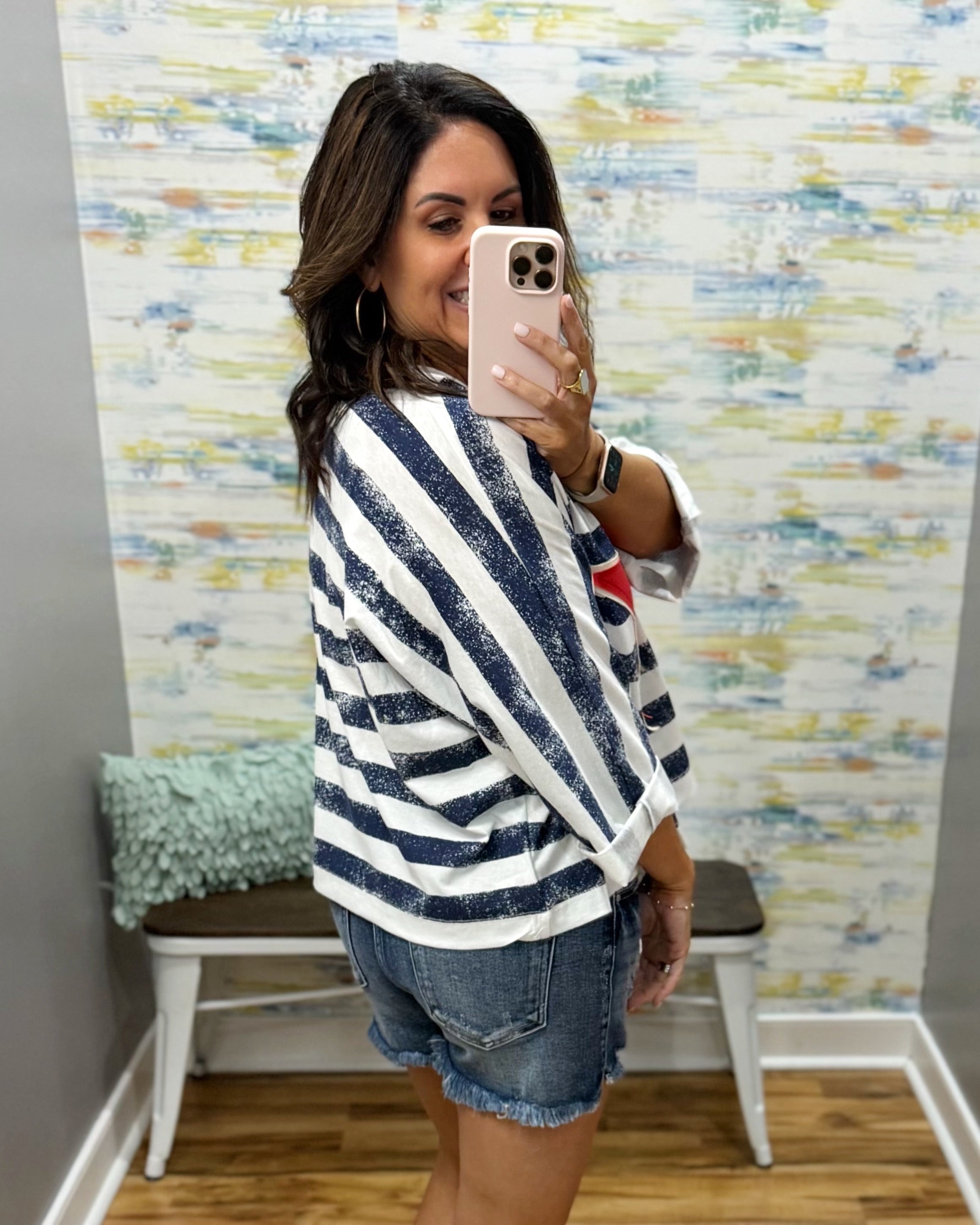 Woman taking a mirror selfie wearing a striped shirt and denim shorts in a room with colorful wall art.