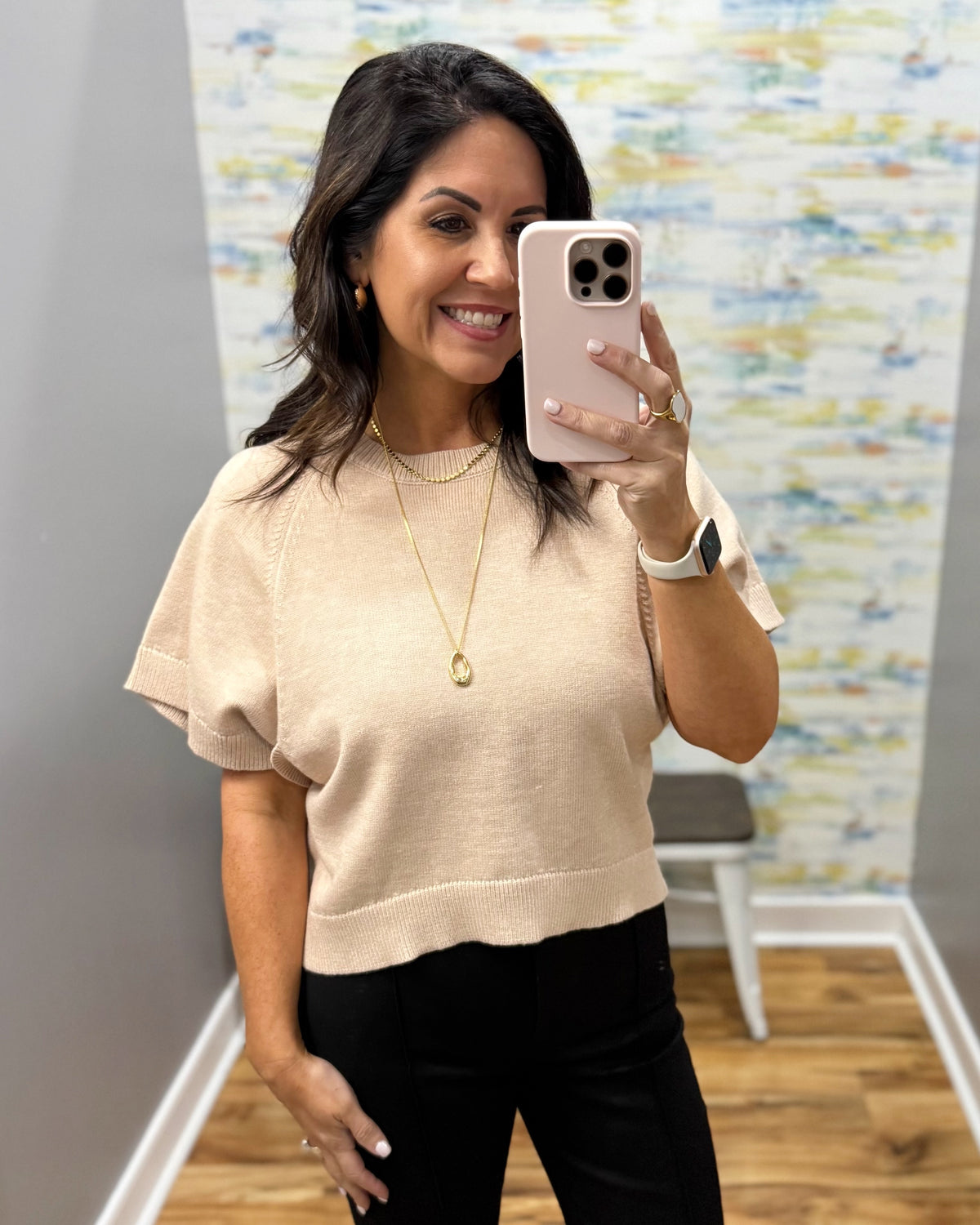 A woman takes a mirror selfie wearing a beige short-sleeve knit top and black pants. She is holding a light pink smartphone, with a gray wall, a colorful abstract wallpaper, and a wooden floor visible in the background.