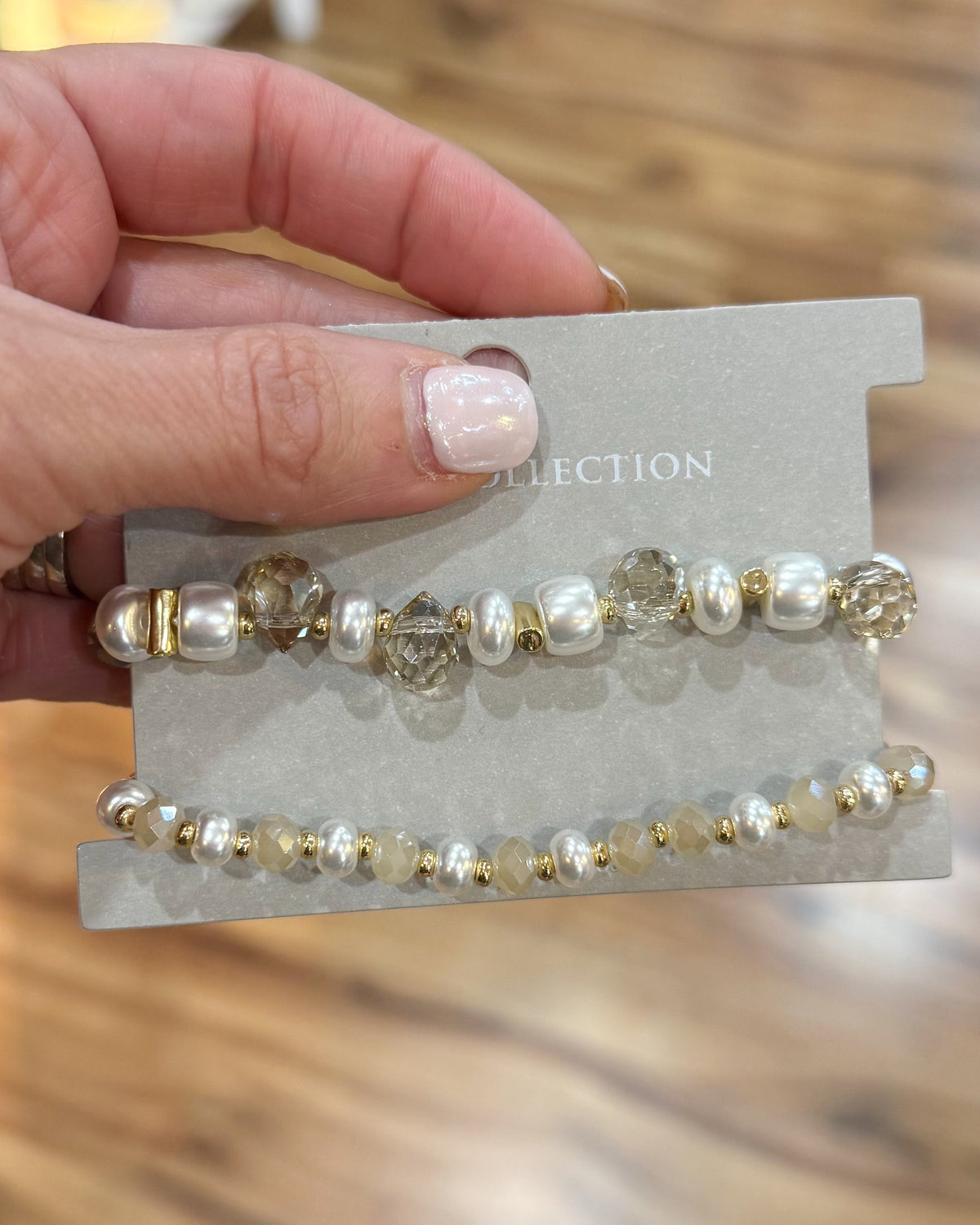 A hand holding a gray jewelry display card featuring two pearl and crystal beaded bracelets. The top bracelet has larger white pearls and faceted clear crystals with gold-toned spacers, while the bottom bracelet has smaller white pearls and gold-toned beads.
