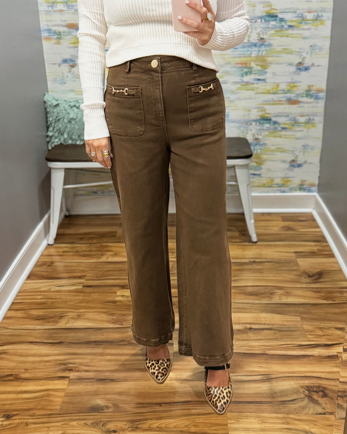 A person wearing brown wide-leg jeans with decorative gold-tone hardware on the back pockets, paired with a cream ribbed sweater and leopard-print pointed flats, standing in an indoor room with wood floors and a bench.