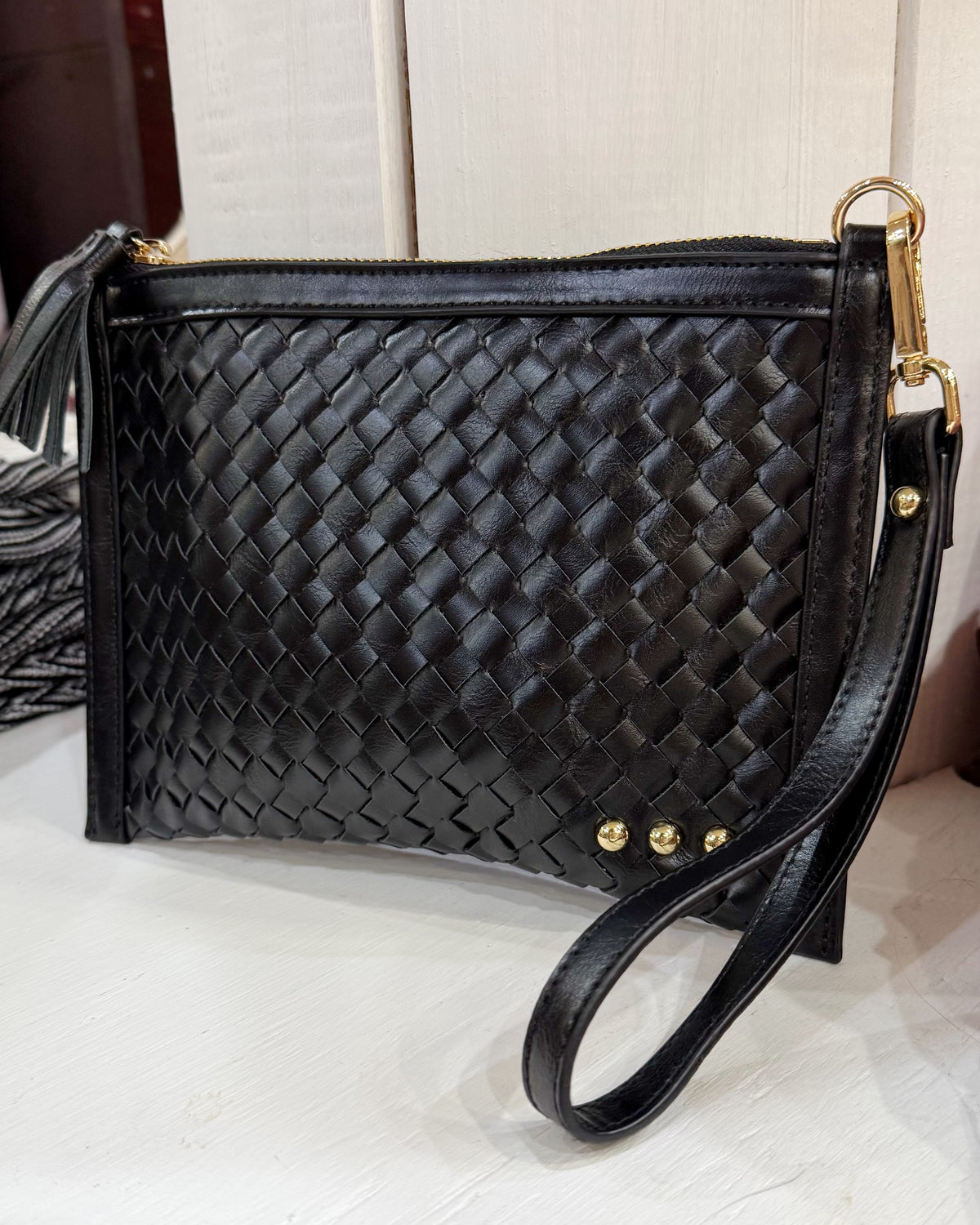 Black woven leather clutch handbag with a wristlet strap, gold-tone hardware, and a tassel detail, displayed on a light surface.