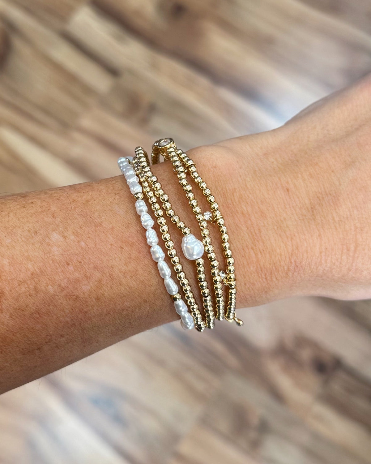 A close-up of a person's wrist wearing three layered bracelets: two gold beaded bracelets and one white pearl beaded bracelet with a teardrop pearl charm, set against a blurred wooden floor background.