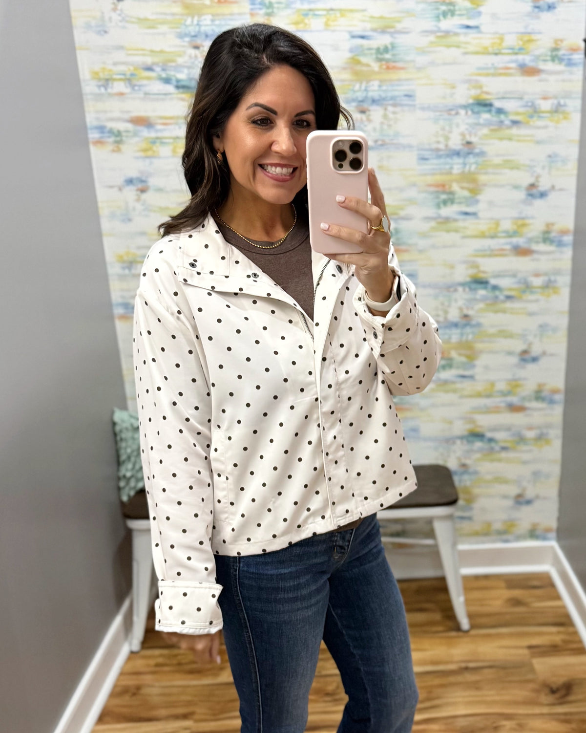 A woman takes a mirror selfie wearing a white long-sleeve button-up shirt with black polka dots, a brown top underneath, and blue jeans in a room with patterned wallpaper and wood floors.