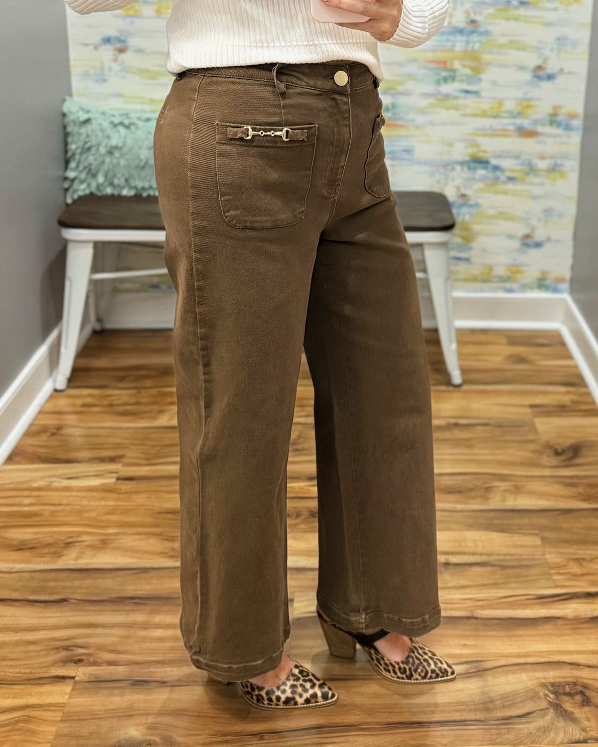 A person wearing brown wide-leg cropped pants with a decorative gold horsebit detail on the back pocket, styled with a cream ribbed sweater and leopard print heels in an indoor setting with a wood floor and bench.