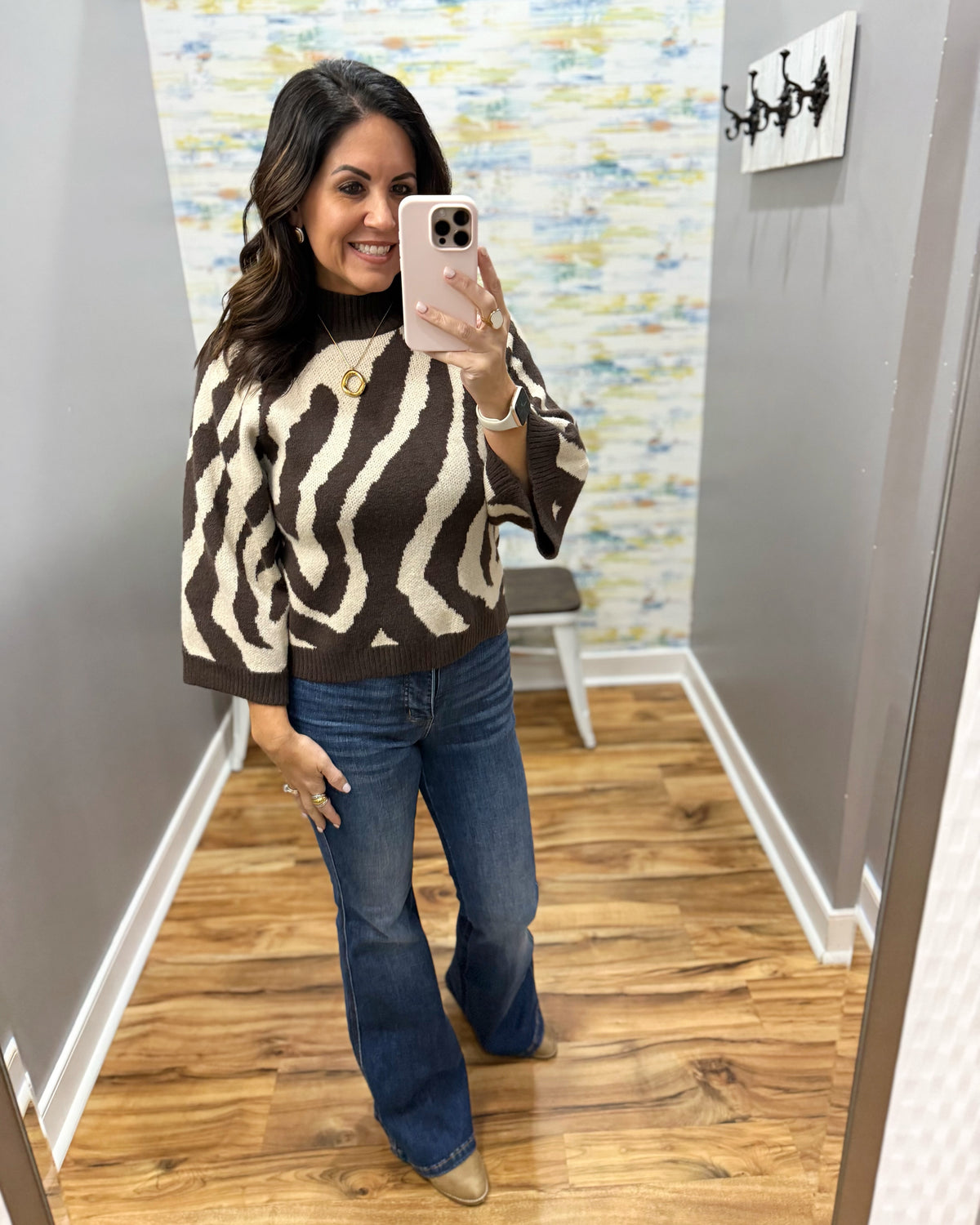 A woman takes a mirror selfie wearing a brown and cream zebra-patterned knit sweater with wide three-quarter sleeves, paired with blue flare jeans and beige ankle boots, in a fitting room with wood floors and gray walls.