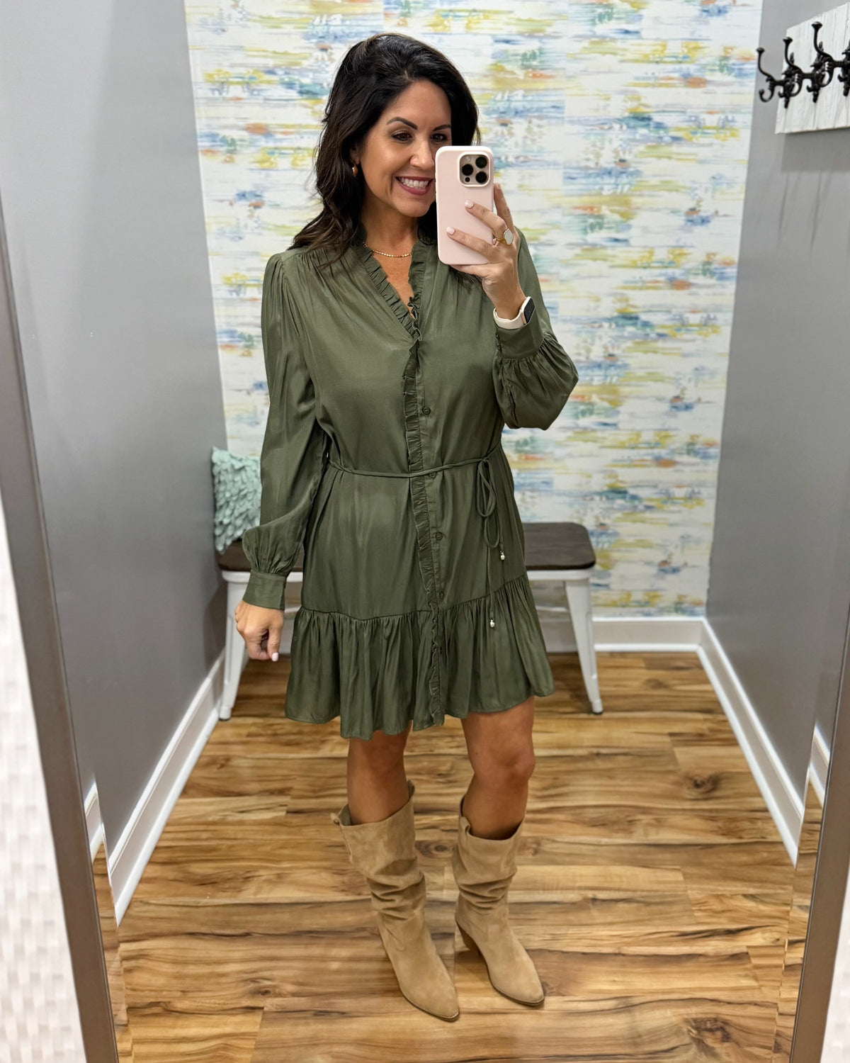 A woman takes a mirror selfie in a fitting room wearing an olive green long-sleeve mini dress with a ruffled neckline and tie waist, paired with tan suede knee-high boots.