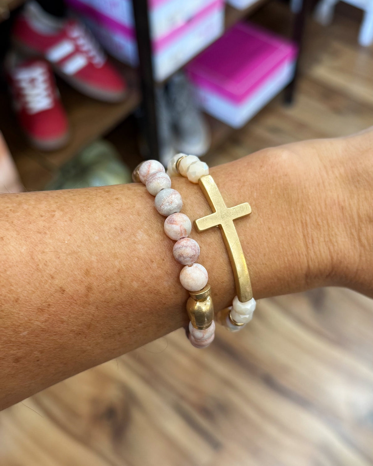 A close-up of a person's wrist wearing two bracelets: one beaded bracelet with pink and white marbled beads and a gold accent bead, and one gold-tone bracelet with a cross charm.