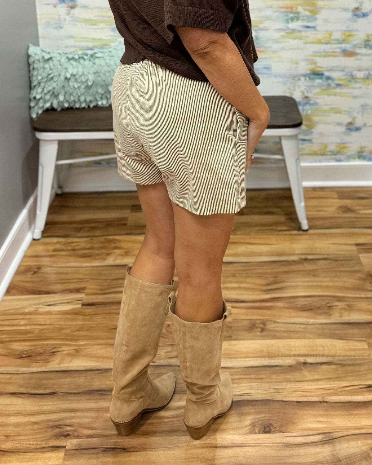 A person shown from the back wearing light beige corduroy shorts and tan knee-high suede boots, standing on a wood floor in an indoor room with a bench and abstract wall art in the background.