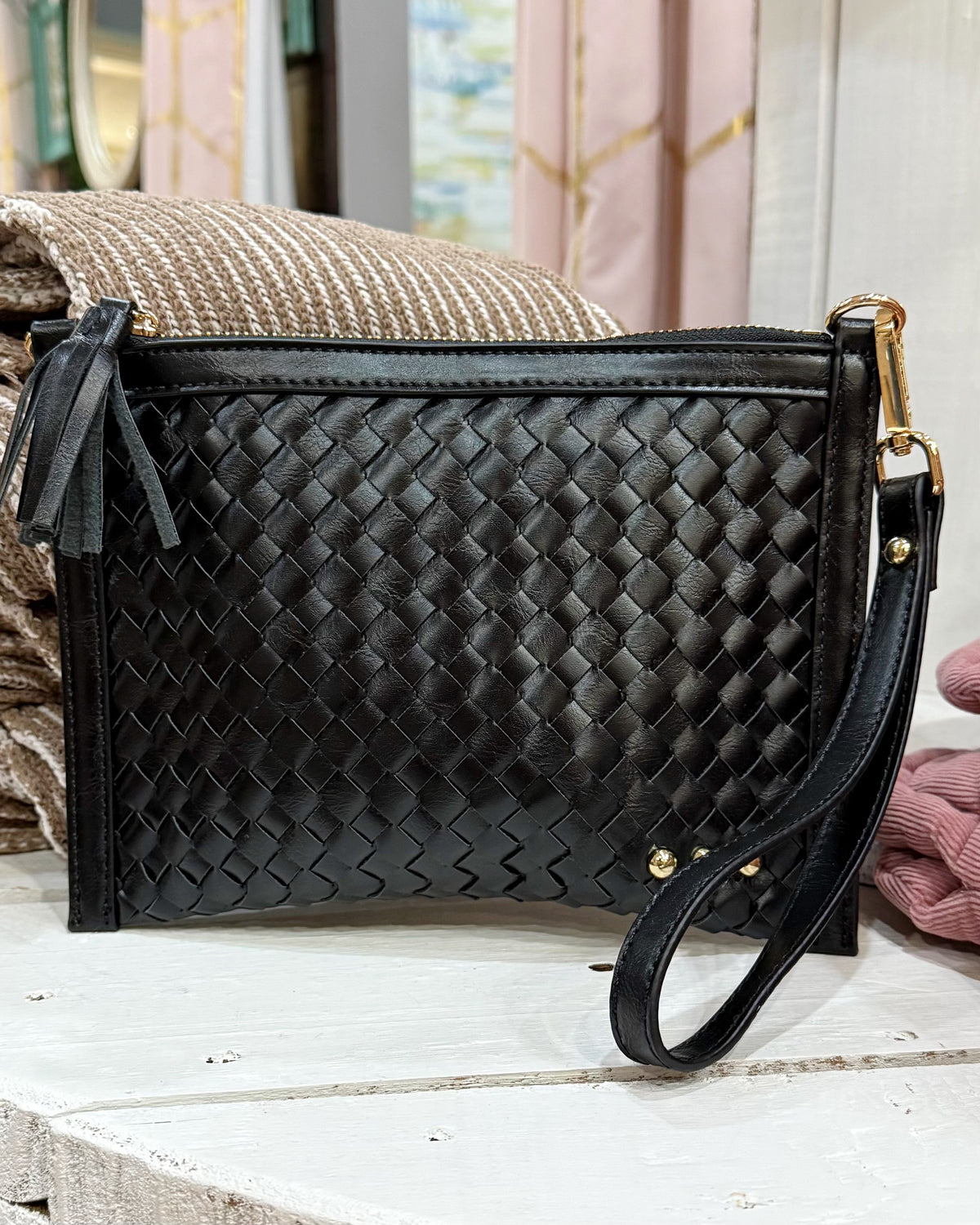 Black woven leather clutch bag with a shoulder strap, gold-tone hardware, and a tassel zipper pull, displayed on a white wooden surface with a woven textile in the background.
