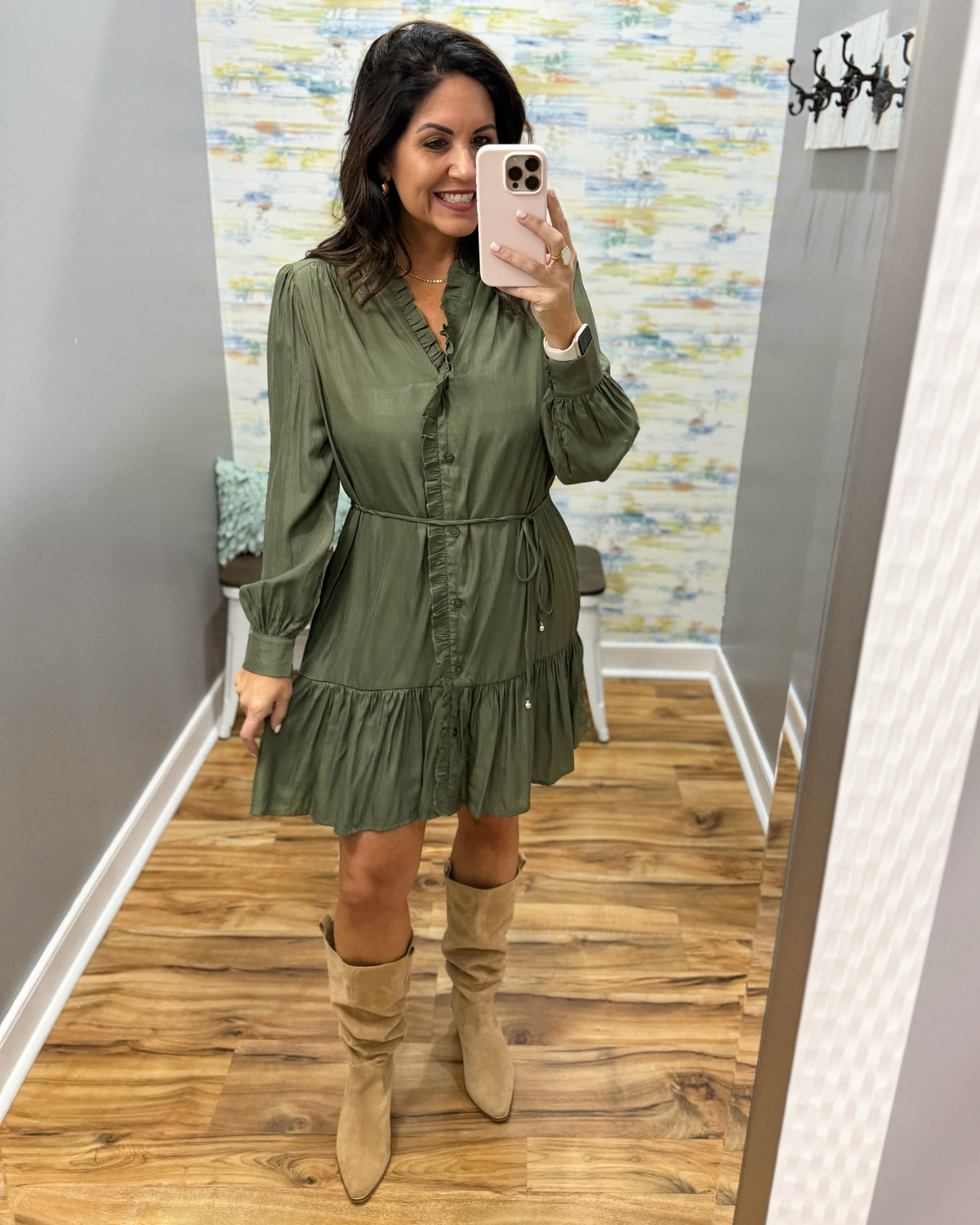 A woman takes a mirror selfie in a fitting room wearing an olive green long-sleeve mini dress with a ruffled hem and beige suede knee-high boots.