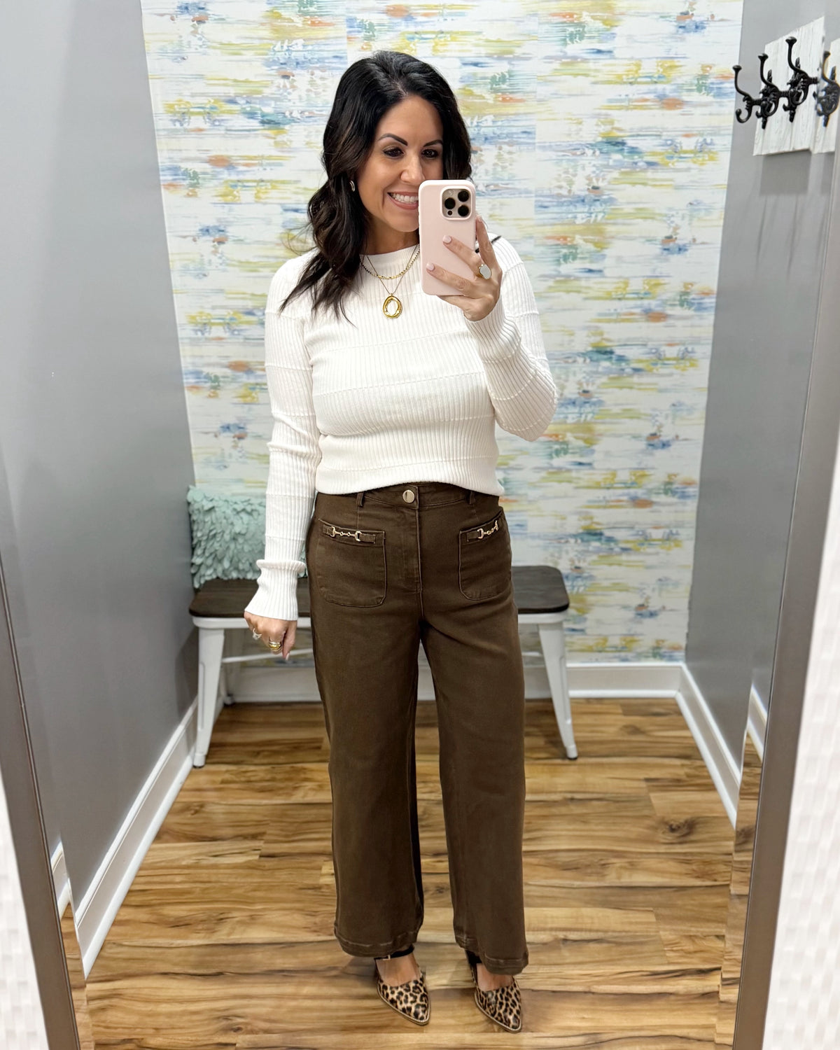 A woman takes a mirror selfie in a fitting room wearing a cream ribbed long-sleeve mock neck sweater and brown wide-leg pants with decorative gold hardware on the front pockets.