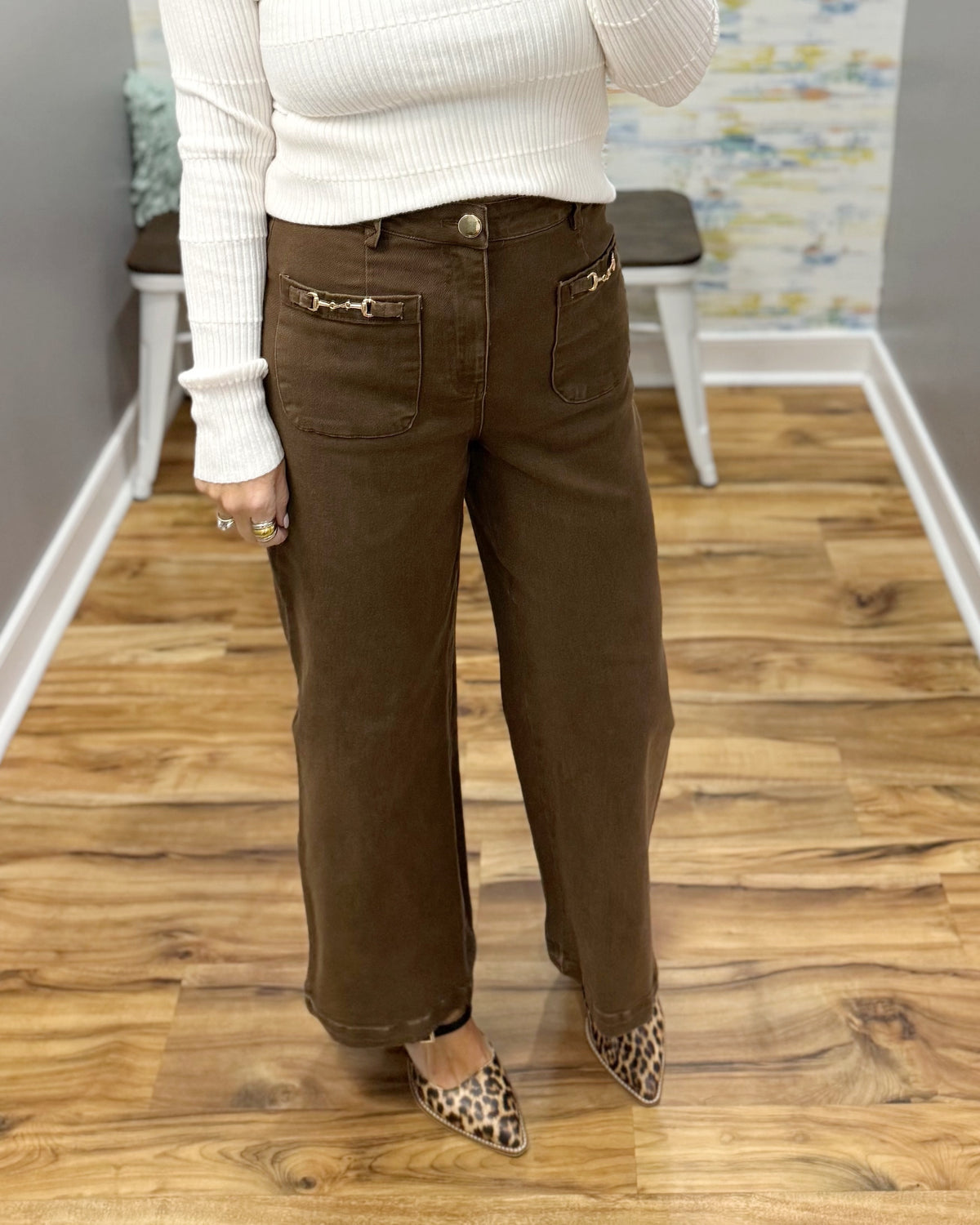 A person wearing brown wide-leg corduroy pants with gold-tone horsebit pocket details, paired with a white ribbed sweater and leopard print flats, standing on a wood floor.