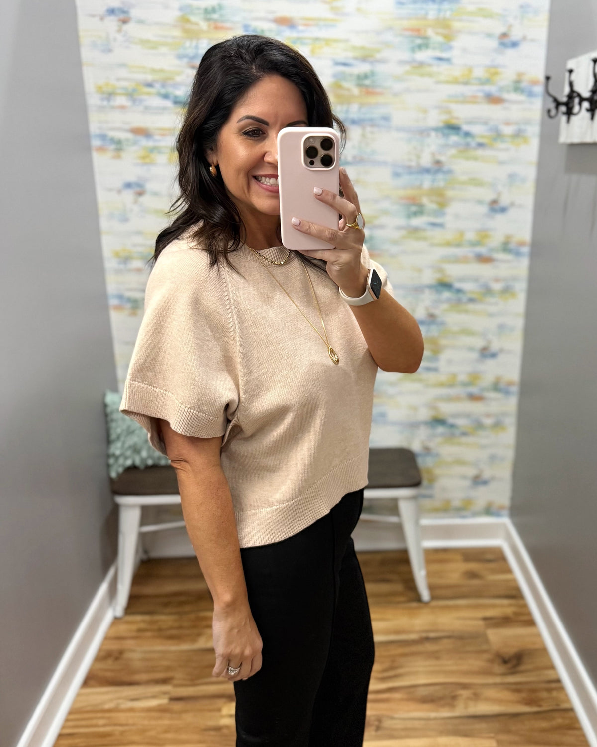 A woman taking a mirror selfie wearing a beige short-sleeve knit top and black pants in a home setting with a wood floor and patterned wallpaper.