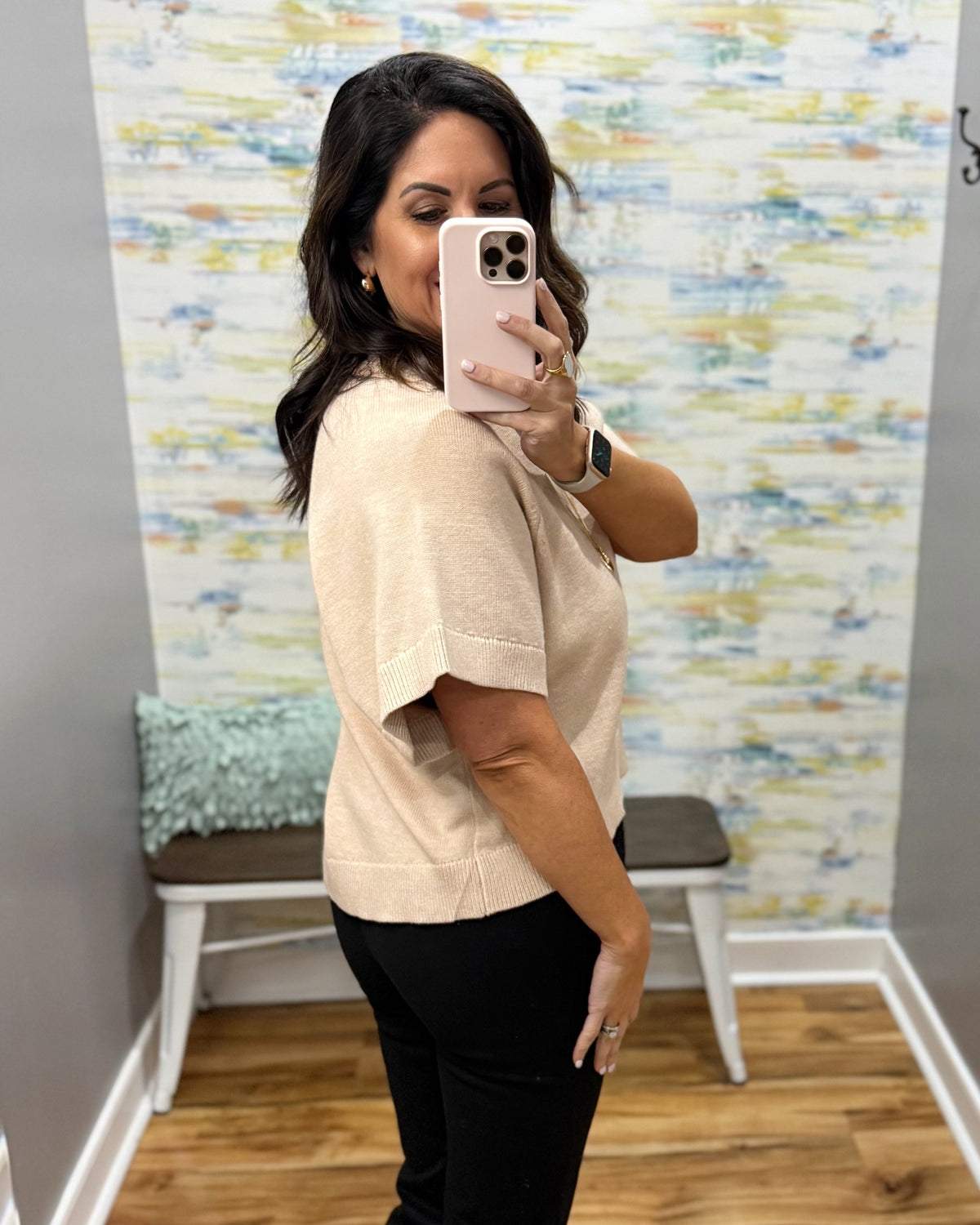 A woman takes a mirror selfie in a fitting room wearing a light beige short-sleeve knit top and black pants. The background features a colorful abstract patterned wall, a wooden bench, and a light wood floor.
