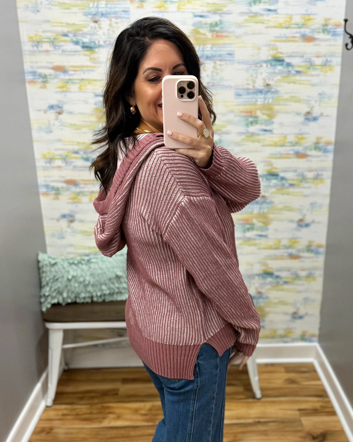 A woman takes a mirror selfie wearing a dusty rose ribbed knit hoodie sweater and blue jeans in a fitting room with a colorful abstract wallpapered wall.