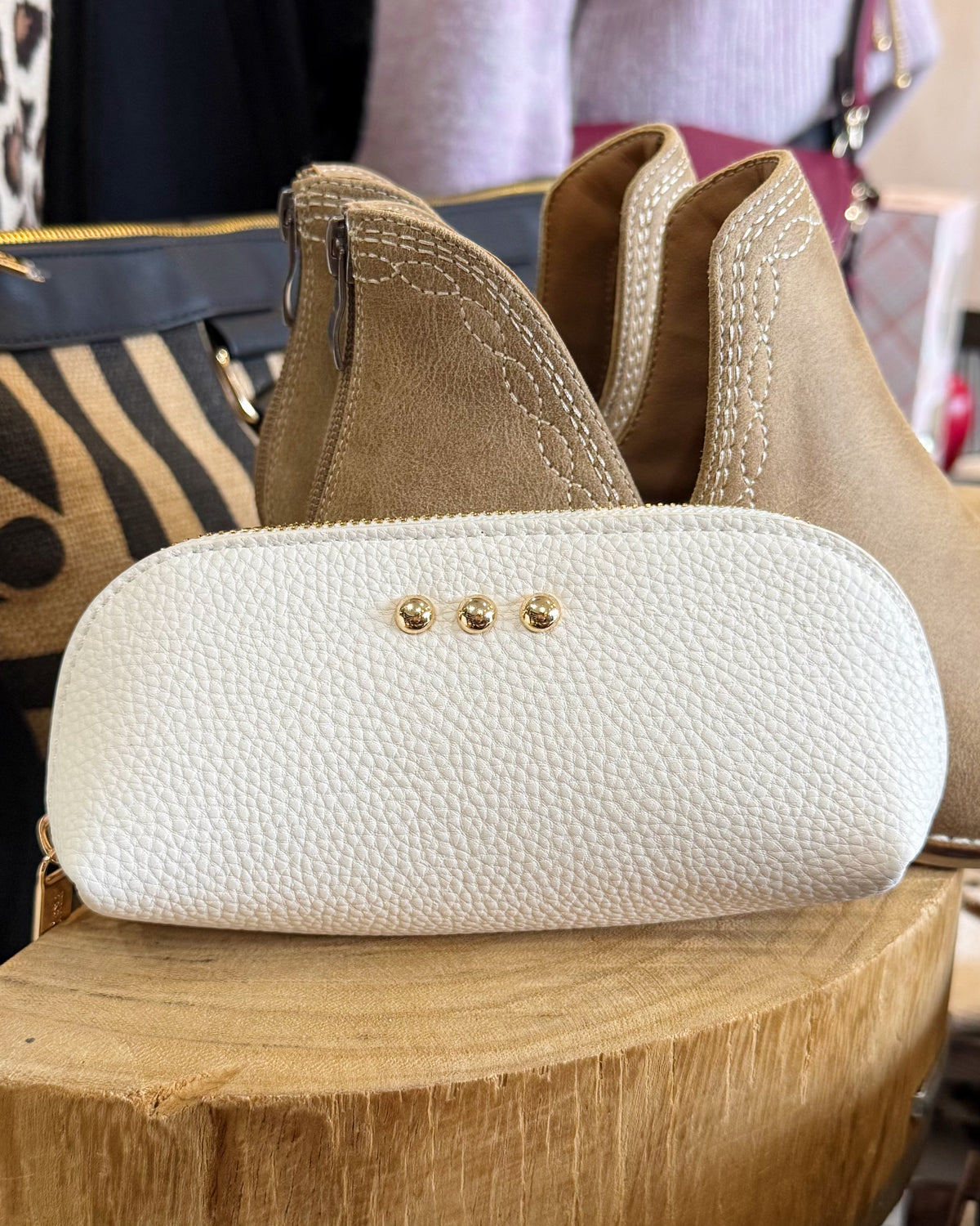 A close-up of a white pebbled leather pouch with three gold-tone studs, displayed on a wooden block with tan suede ankle boots and a patterned handbag in the background.