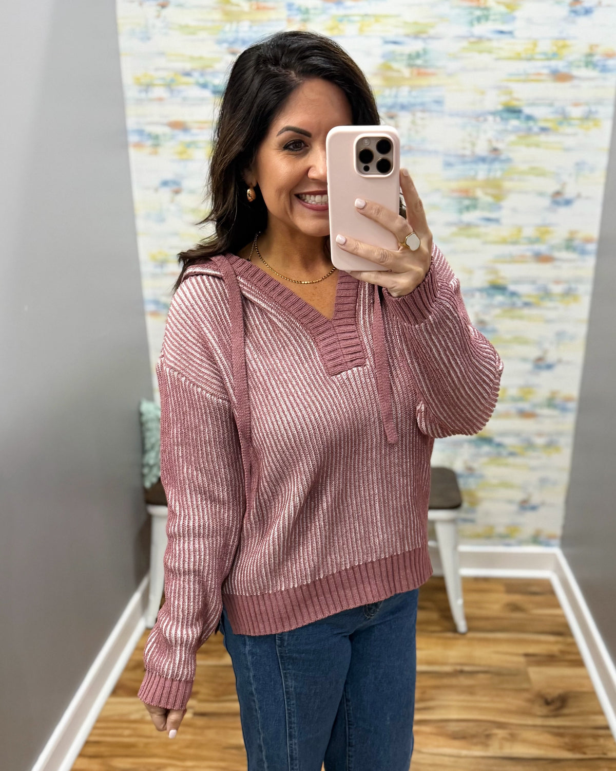 A woman takes a mirror selfie wearing a dusty rose pink ribbed knit hoodie sweater with a V-neck and drawstring hood, paired with blue jeans in an indoor setting.