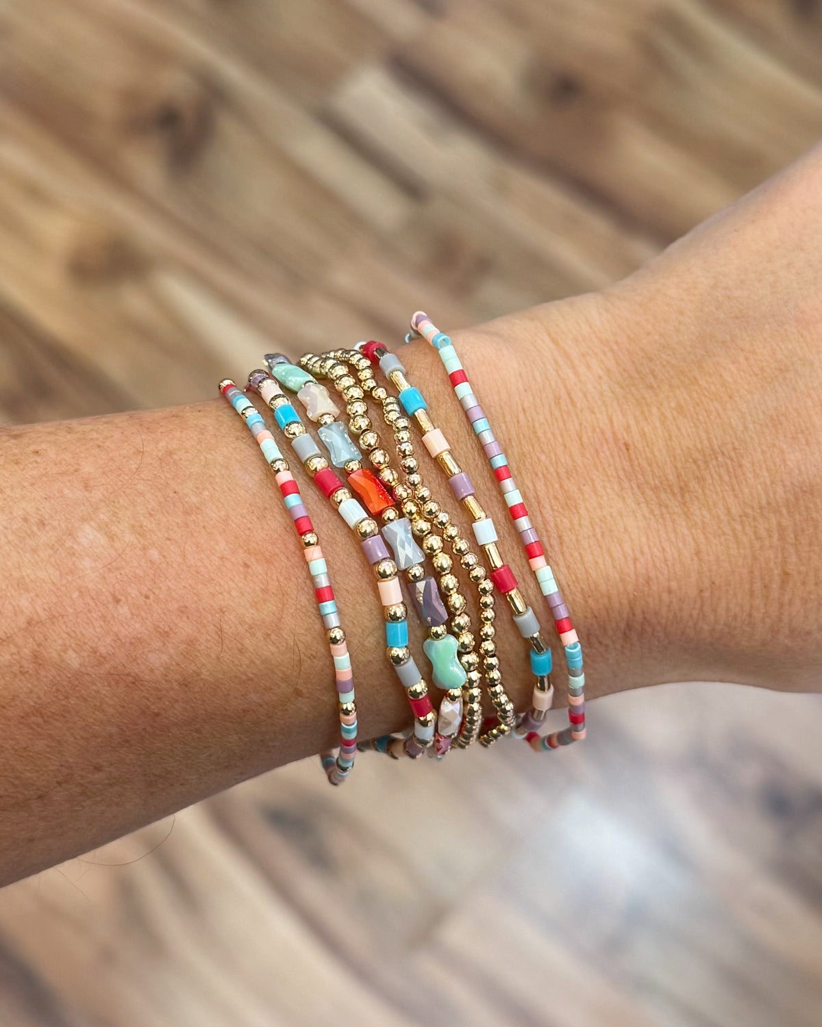 A close-up of a person's wrist wearing a stack of colorful beaded stretch bracelets with gold-toned spacer beads, set against a blurred wooden floor background.