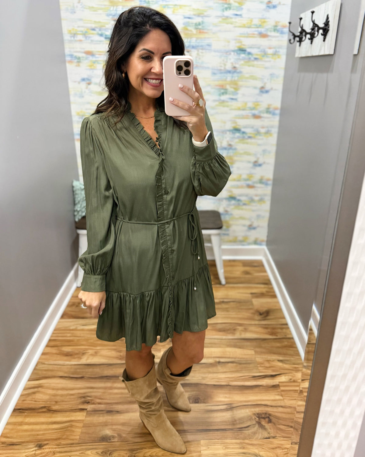 A woman takes a mirror selfie wearing an olive green long-sleeve mini dress with ruffles and beige suede boots in a home setting with wood floors and gray walls.