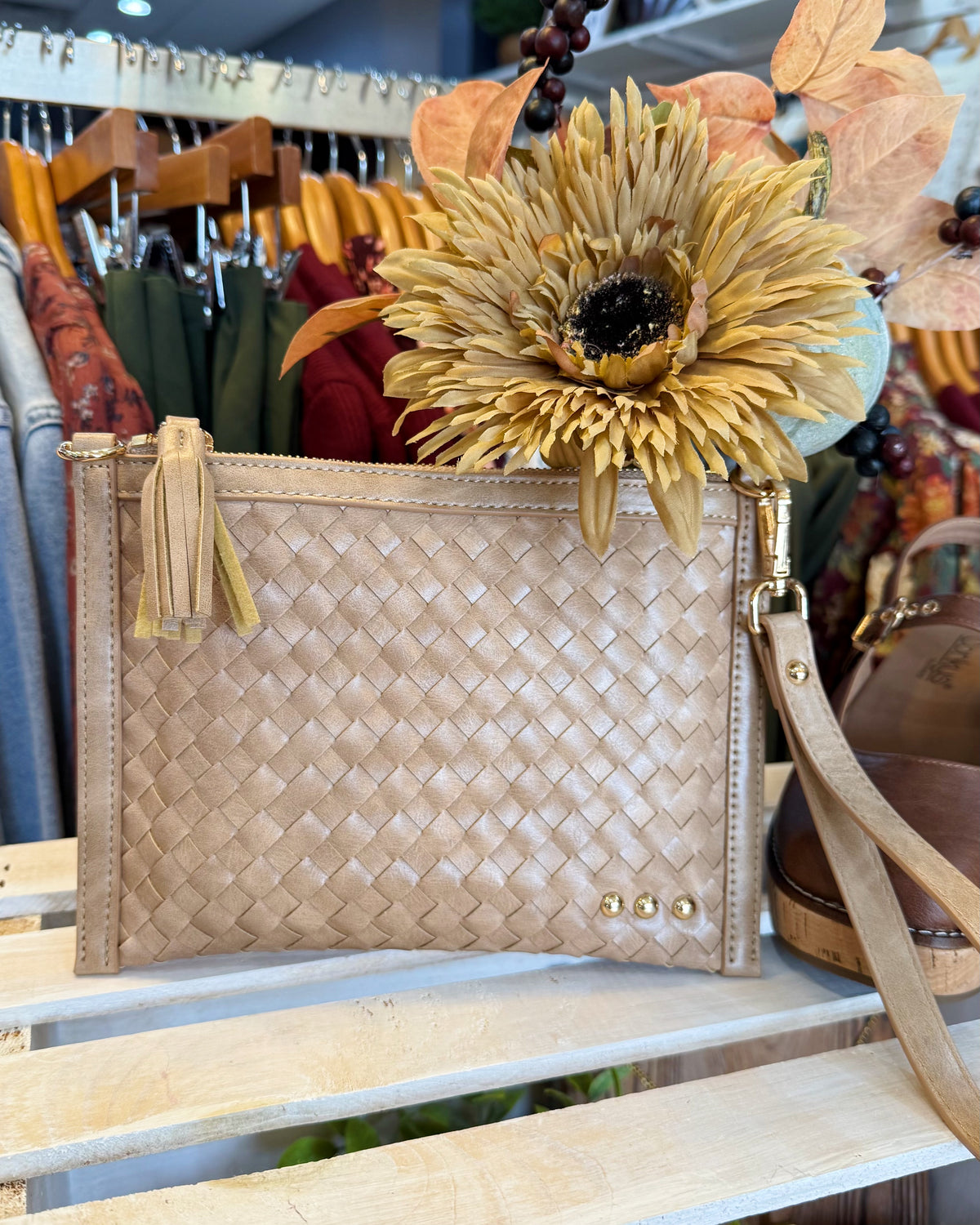 A woven beige clutch bag with a tassel zipper pull and gold-tone hardware, styled on a wooden shelf in a boutique with dried flowers and clothing racks in the background.
