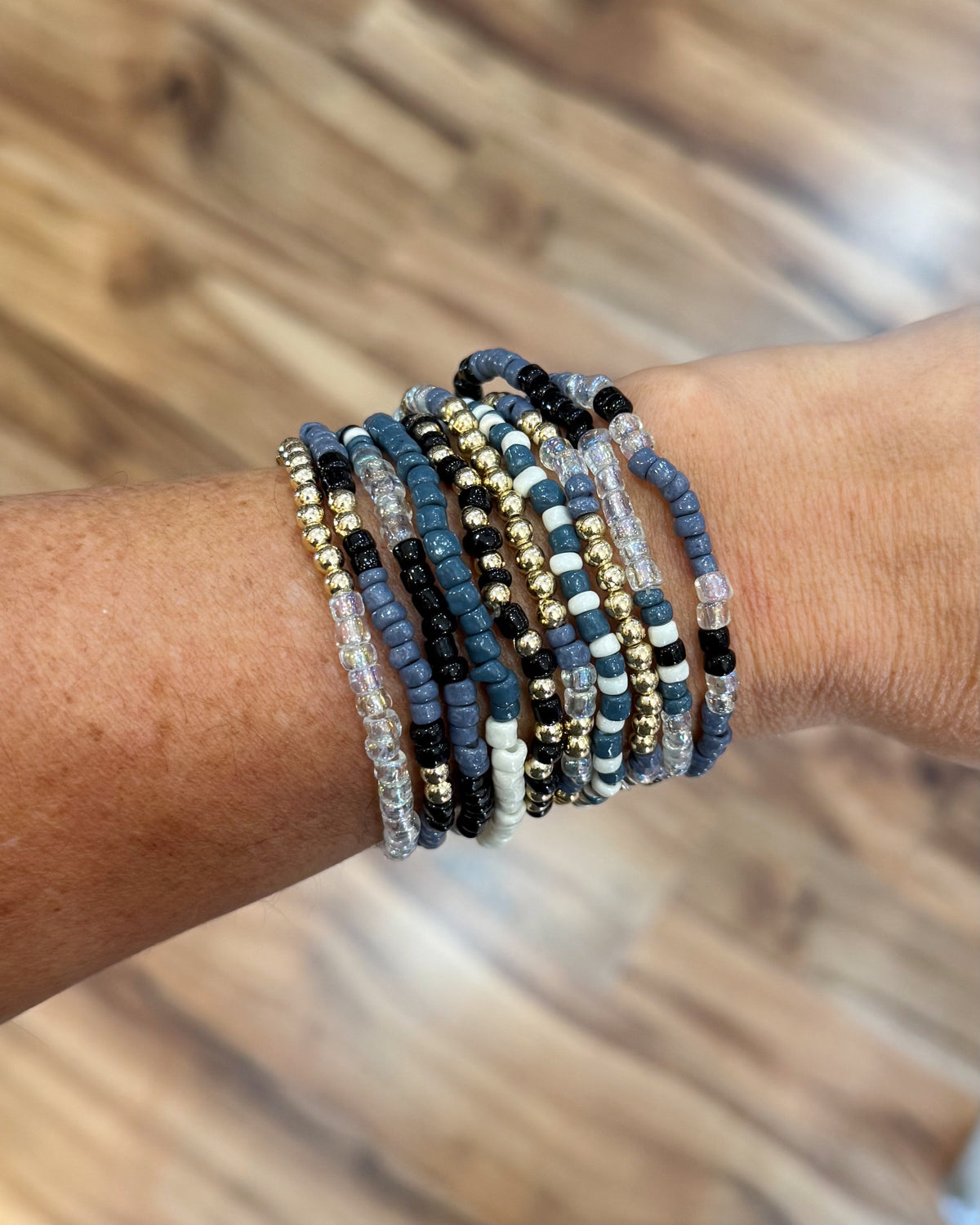 A stack of beaded stretch bracelets worn on a wrist, featuring blue, black, white, and gold beads, with a blurred wood floor in the background.