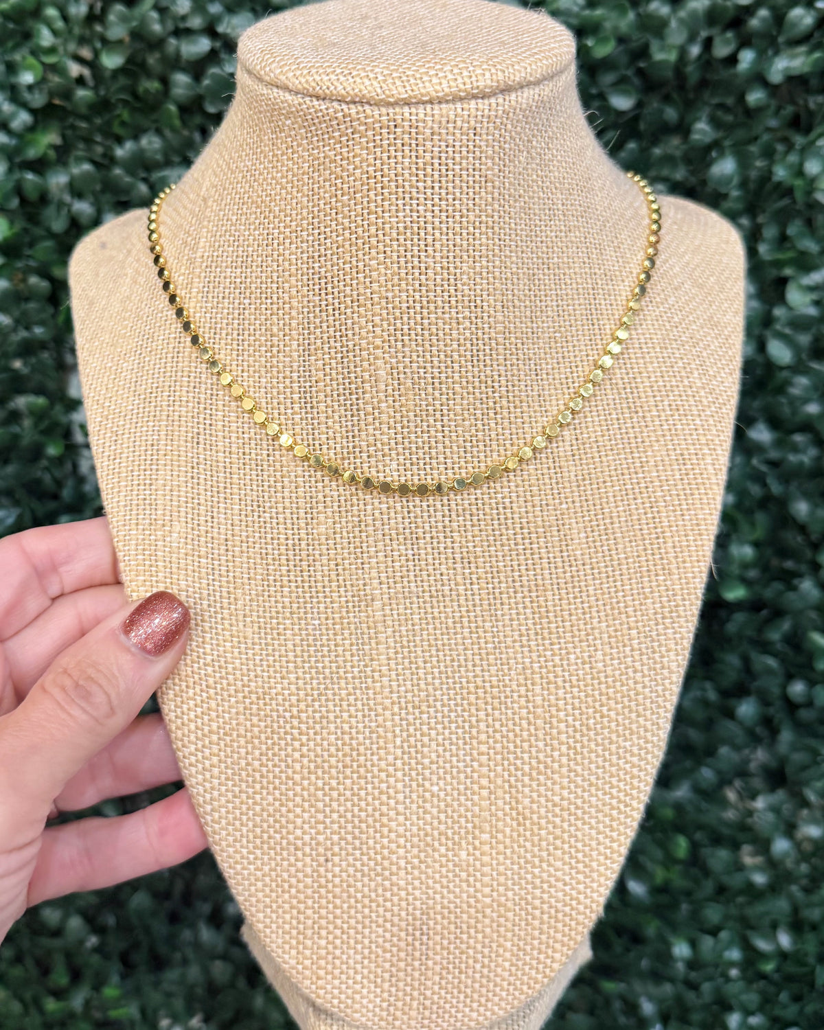 A gold chain necklace displayed on a beige fabric jewelry bust, with a hand holding the left side of the bust. The background is a dark green leafy hedge.