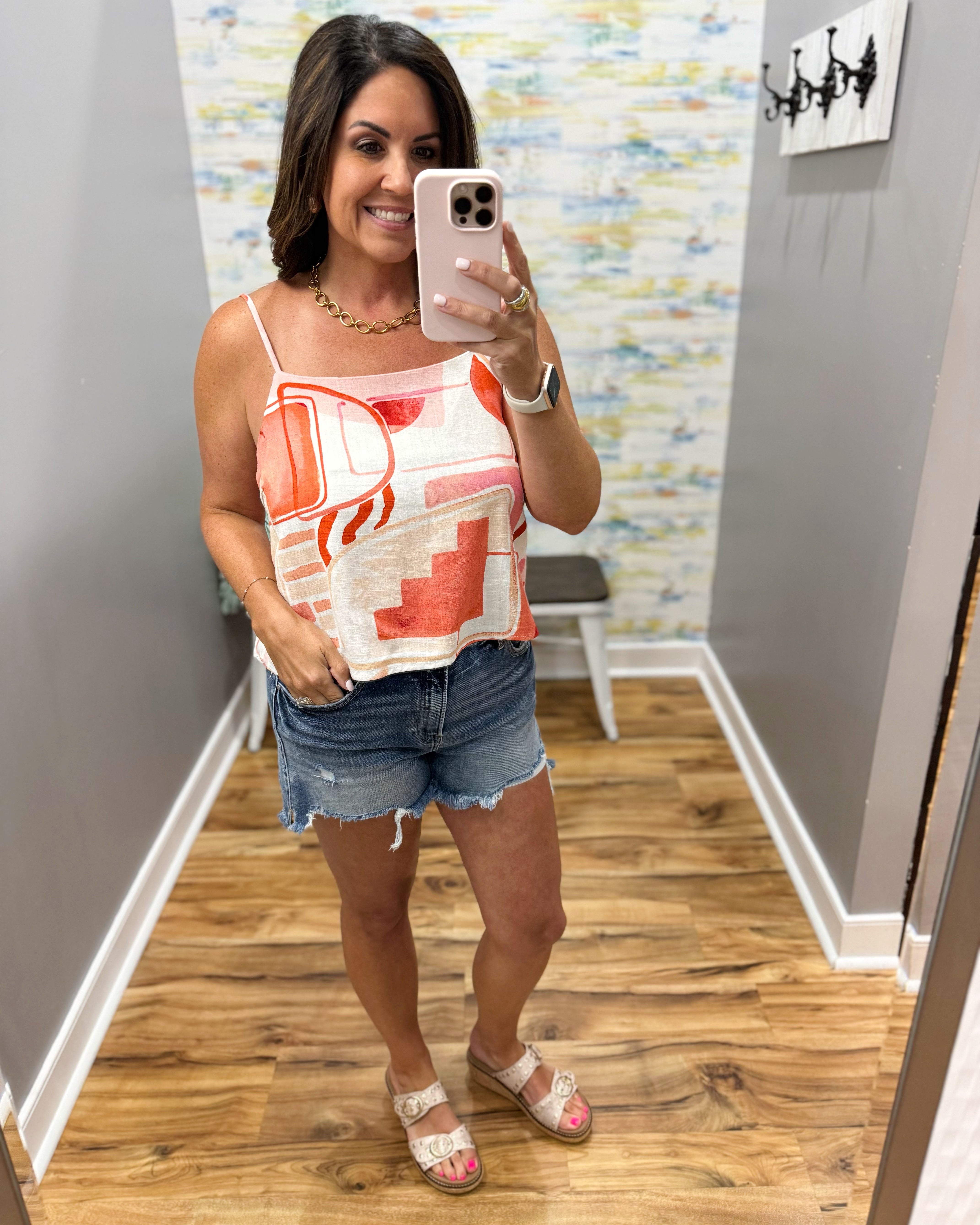 Woman taking a mirror selfie wearing a sleeveless top and denim shorts in a room with a colorful wall.
