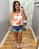 Woman taking a mirror selfie wearing a colorful tank top and denim shorts in a room with patterned wallpaper.