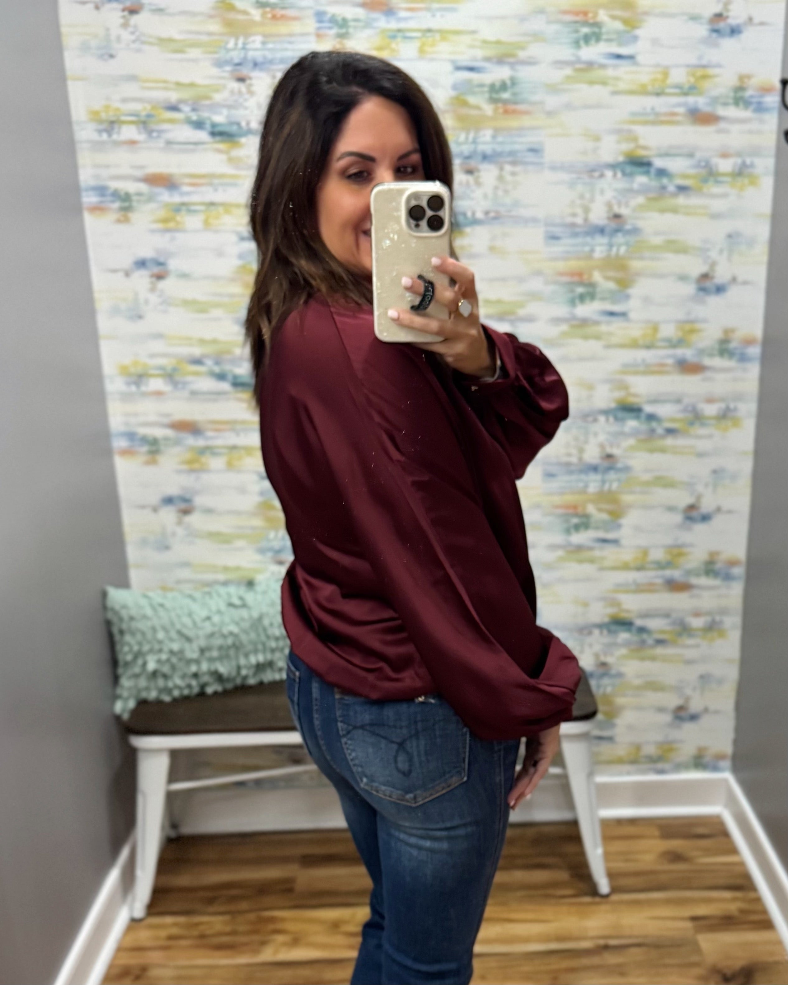 A woman takes a mirror selfie wearing a shiny burgundy satin long-sleeve blouse and blue jeans in a room with patterned wallpaper and a small bench.