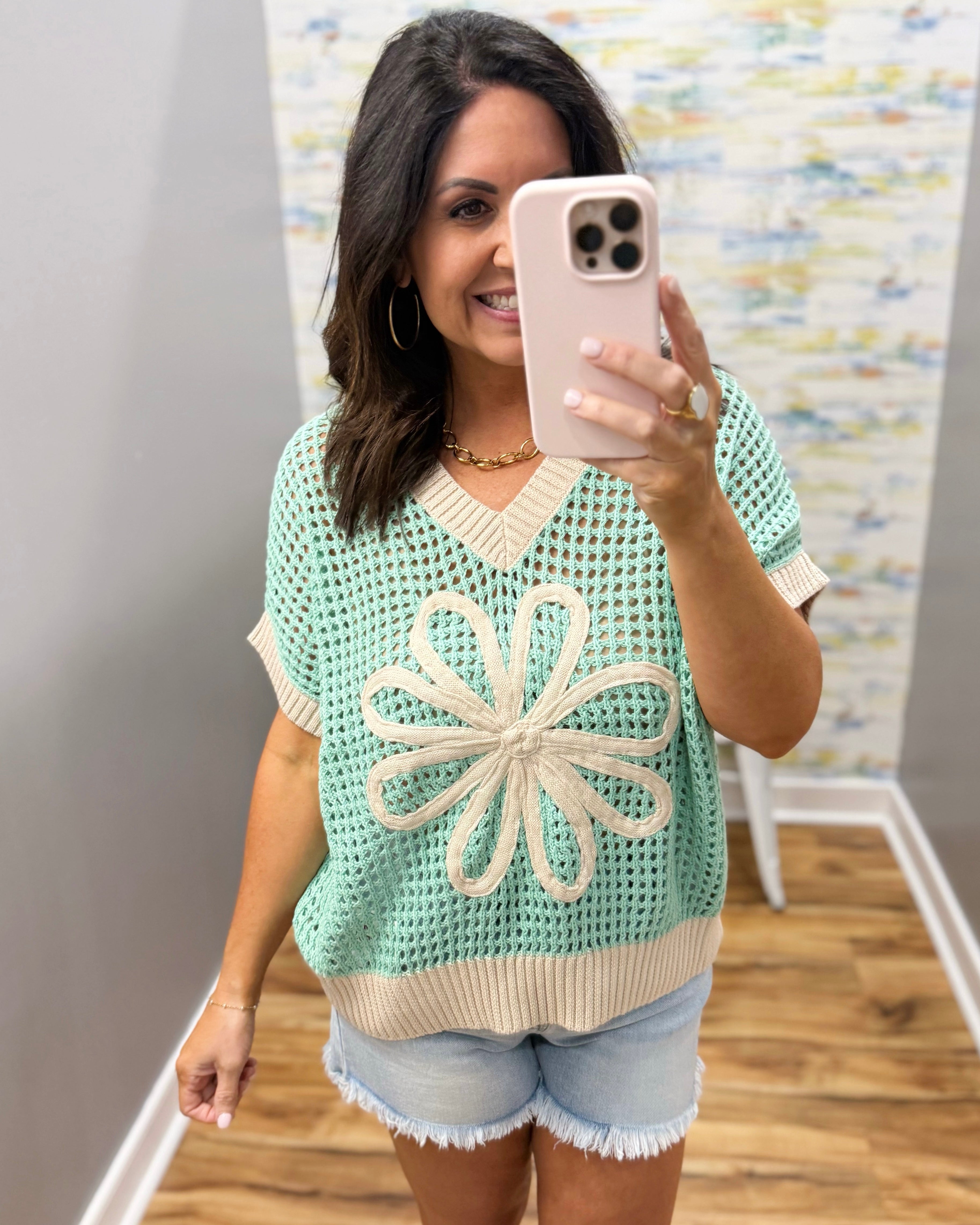Woman taking a mirror selfie wearing a light green sweater with a flower design.