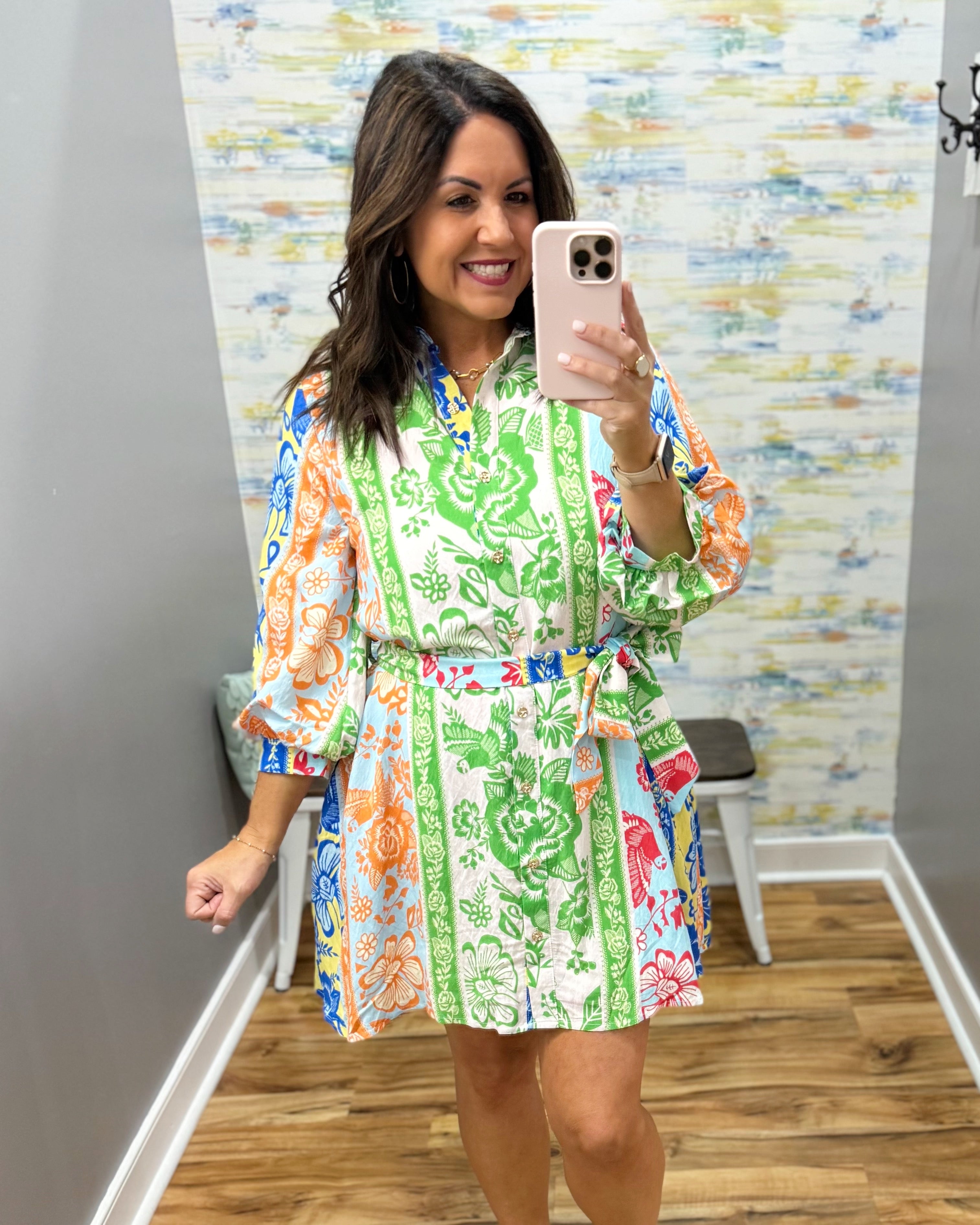 Woman taking a mirror selfie wearing a colorful floral dress in a room with a patterned wall.