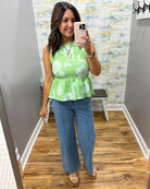 Woman taking a mirror selfie wearing a green floral top and blue jeans in a room with patterned wallpaper.