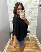 Woman taking a mirror selfie wearing a black top and denim shorts in a room with colorful wall art.