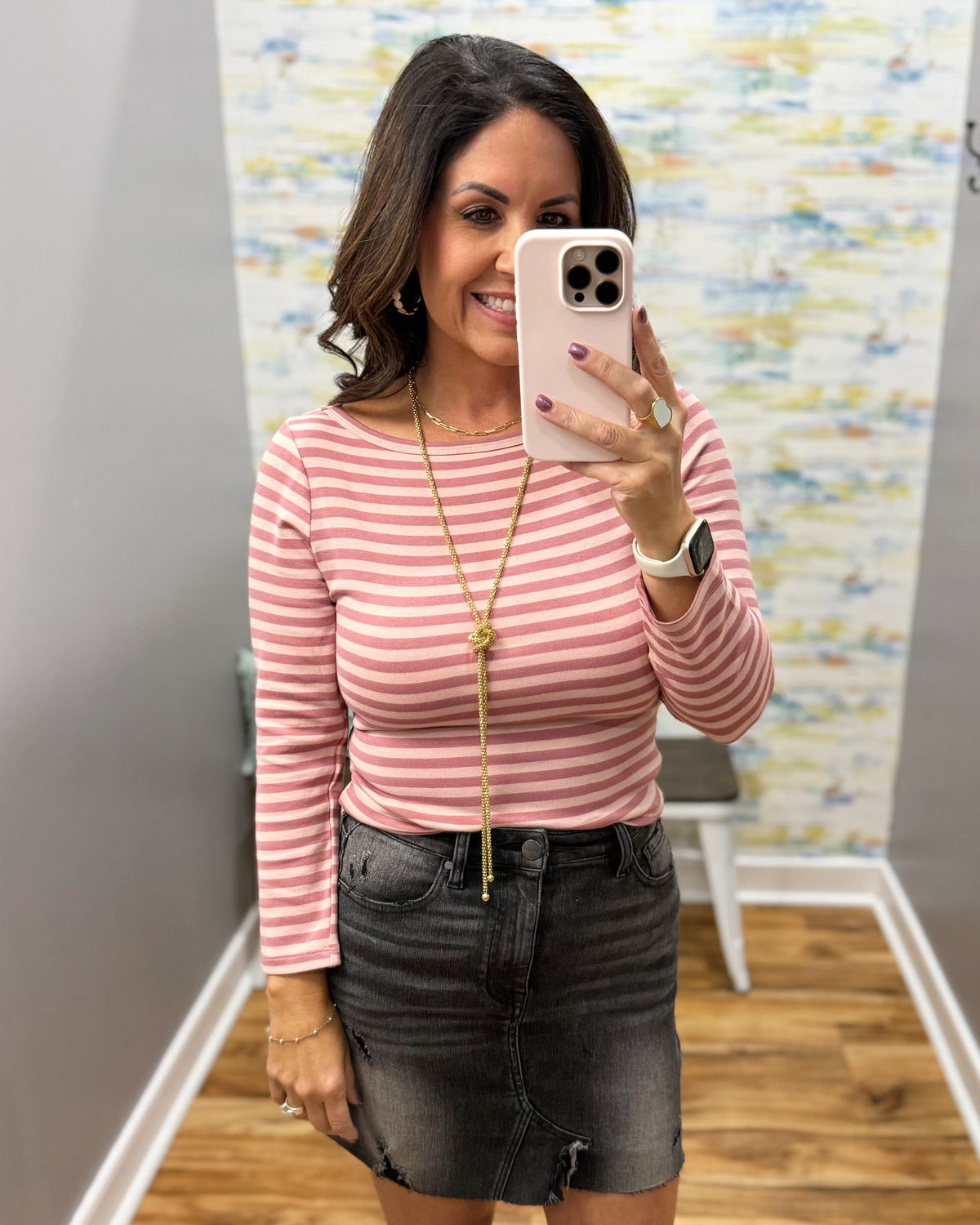 A woman taking a mirror selfie in a fitting room wearing a ginger and mauve striped long-sleeve top and a dark gray denim mini skirt.